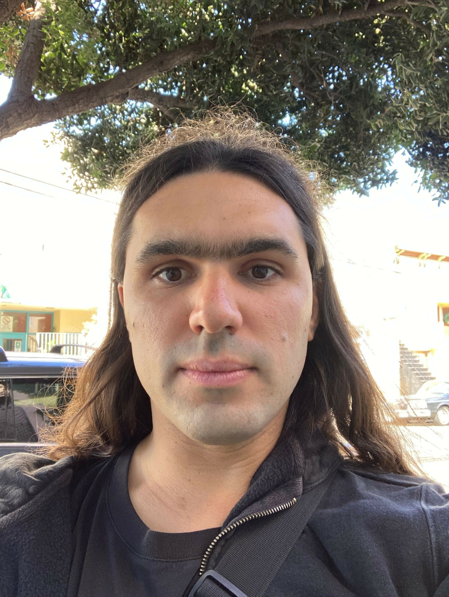 Young person with long hair outdoors, trees and buildings in background, wearing dark jacket, looking at camera.