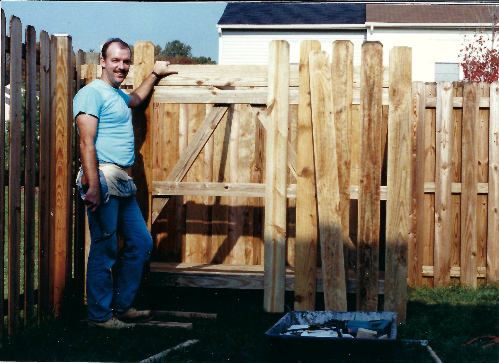 Man standing next to a wooden fence and gate under construction outdoors, with tools and materials nearby.