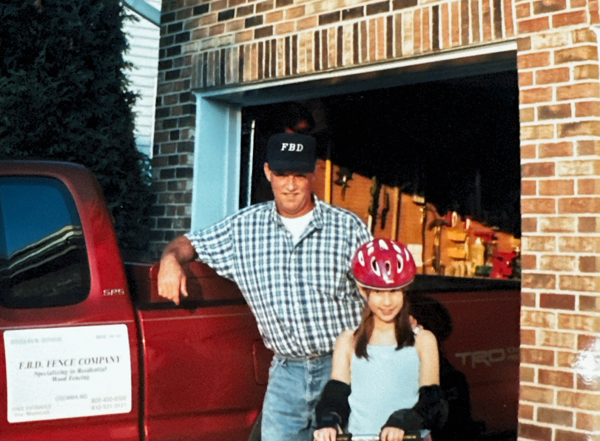 A man and a girl stand outside a brick building, with the man leaning on a red truck and the girl wearing a helmet.