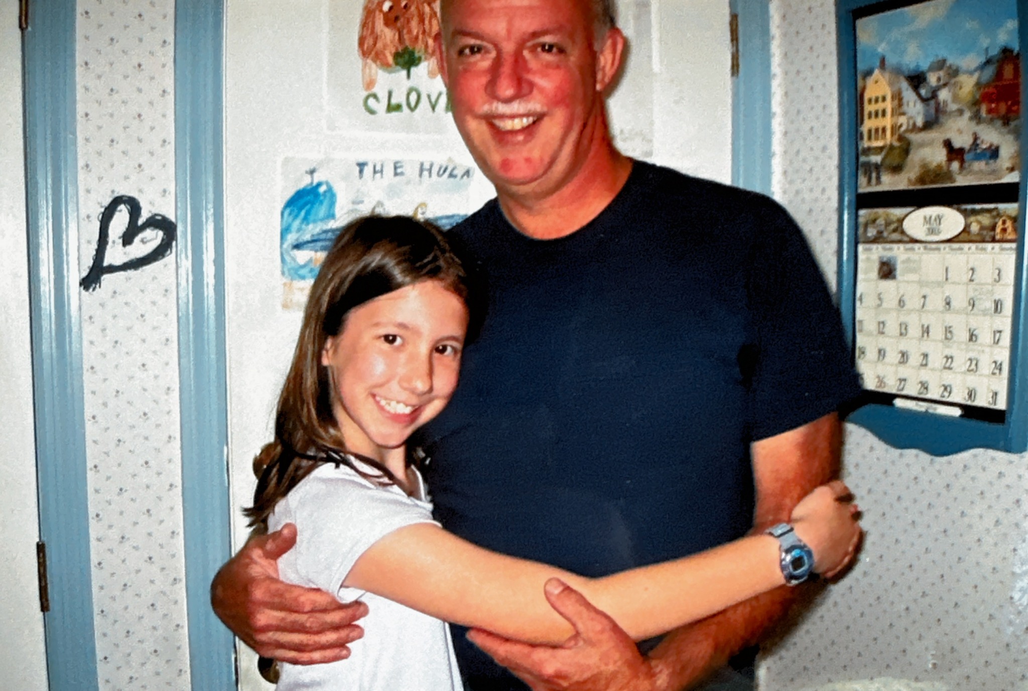 A man and a girl hugging and smiling indoors, with posters and a calendar on the wall behind them.