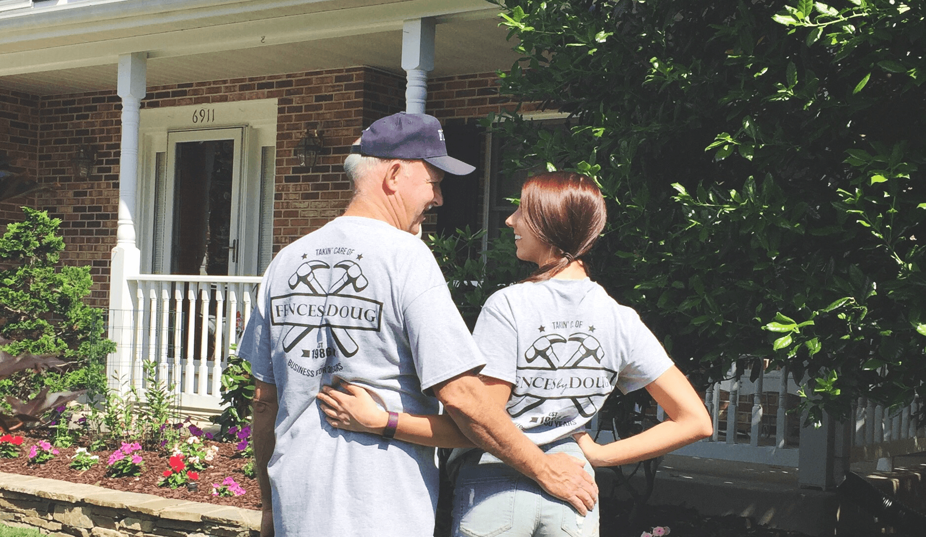 Two people wearing matching t-shirts standing outdoors near a house and trees.