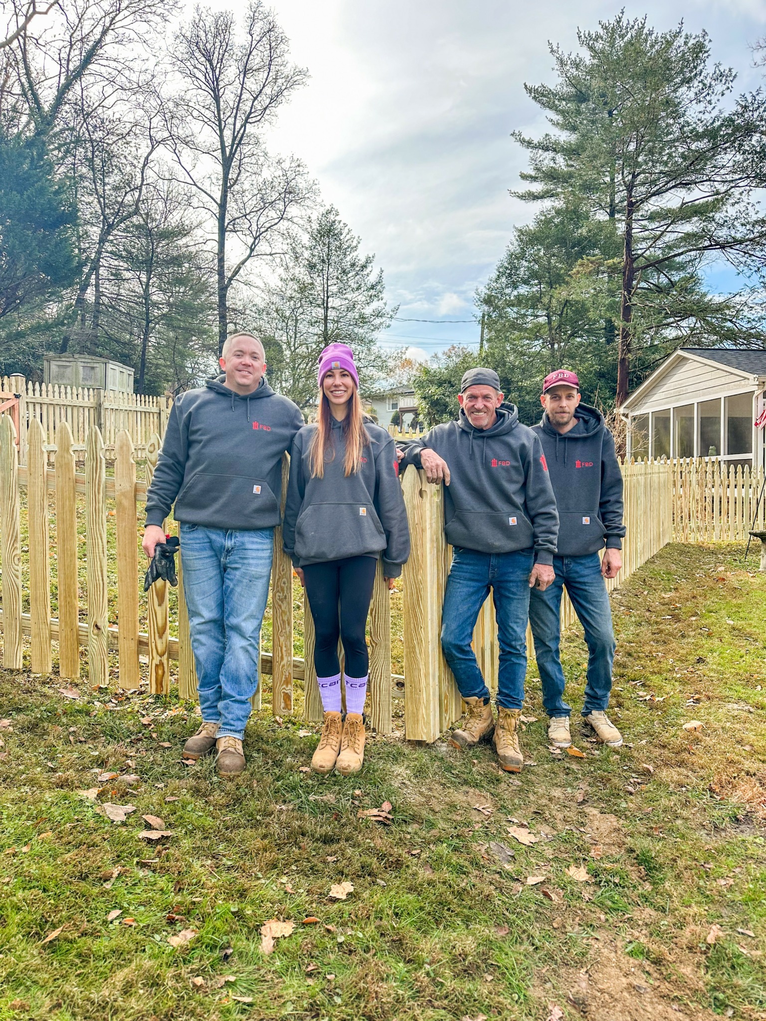 Five people standing outdoors on grass with trees and a wooden fence in the background.