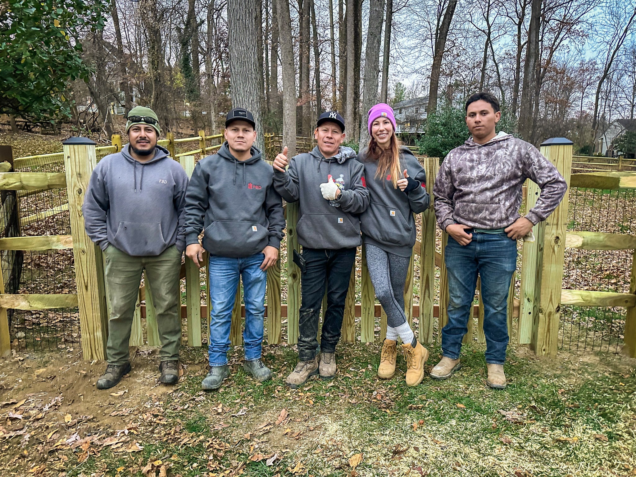 Five people standing outdoors in front of a wooden fence, trees in the background, some with hands in pockets or arms around each other.