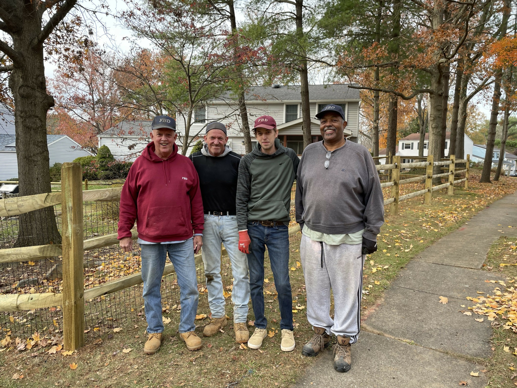 Four people standing on a sidewalk in a park with trees and a wooden fence, autumn leaves on the ground.