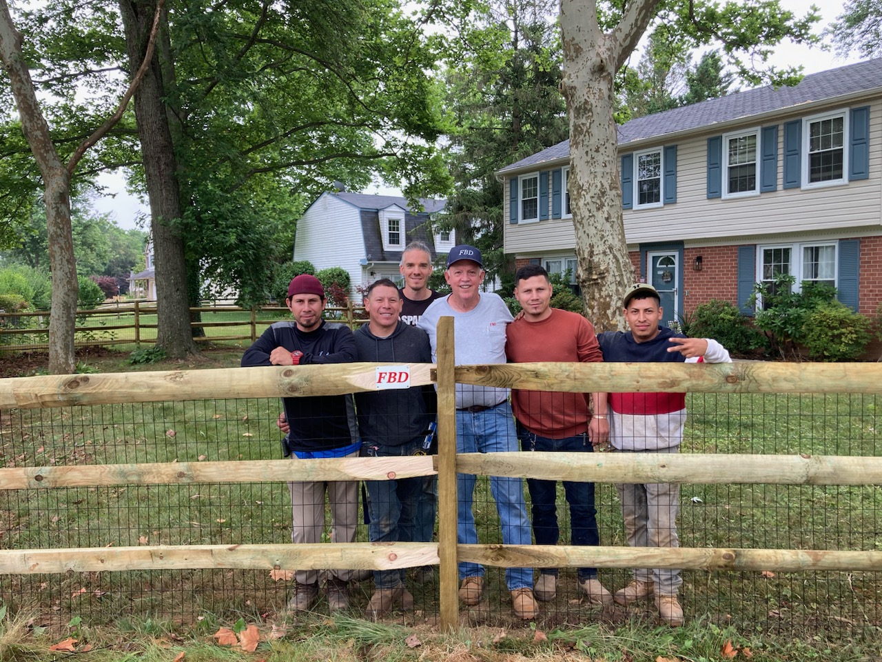 Five people standing outdoors behind a wooden fence in front of a house and trees.