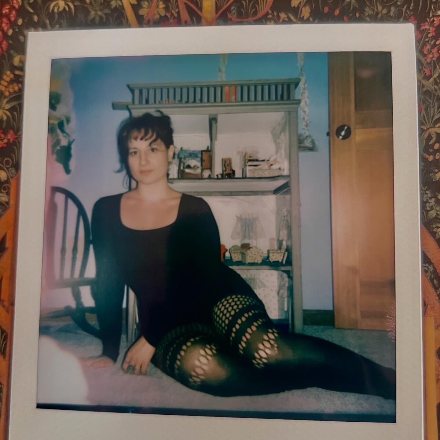 Person with short dark hair sitting on bed, wearing black top and patterned tights, in room with shelves and door.