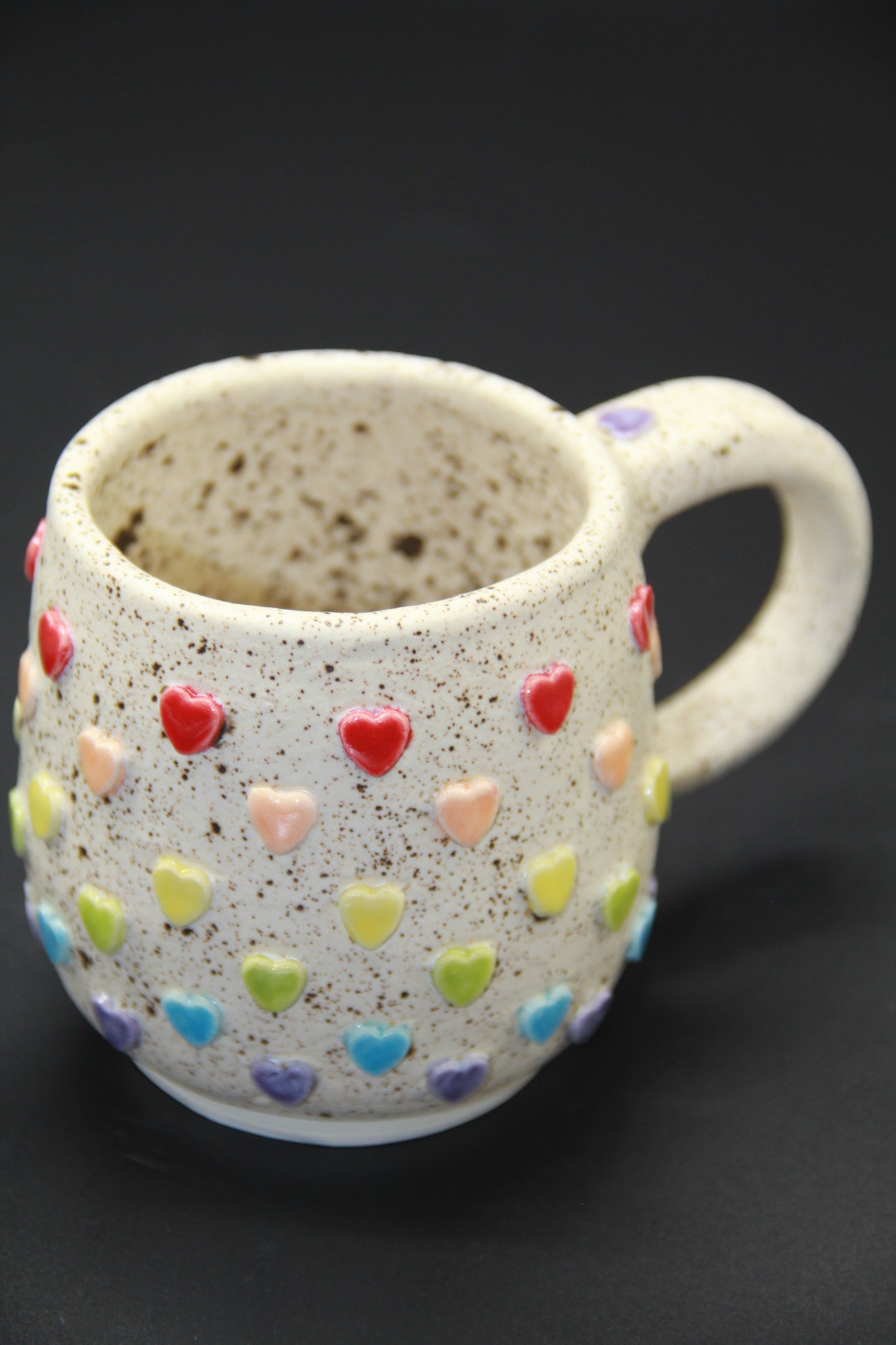 Speckled ceramic mug with colorful heart patterns on a black background.