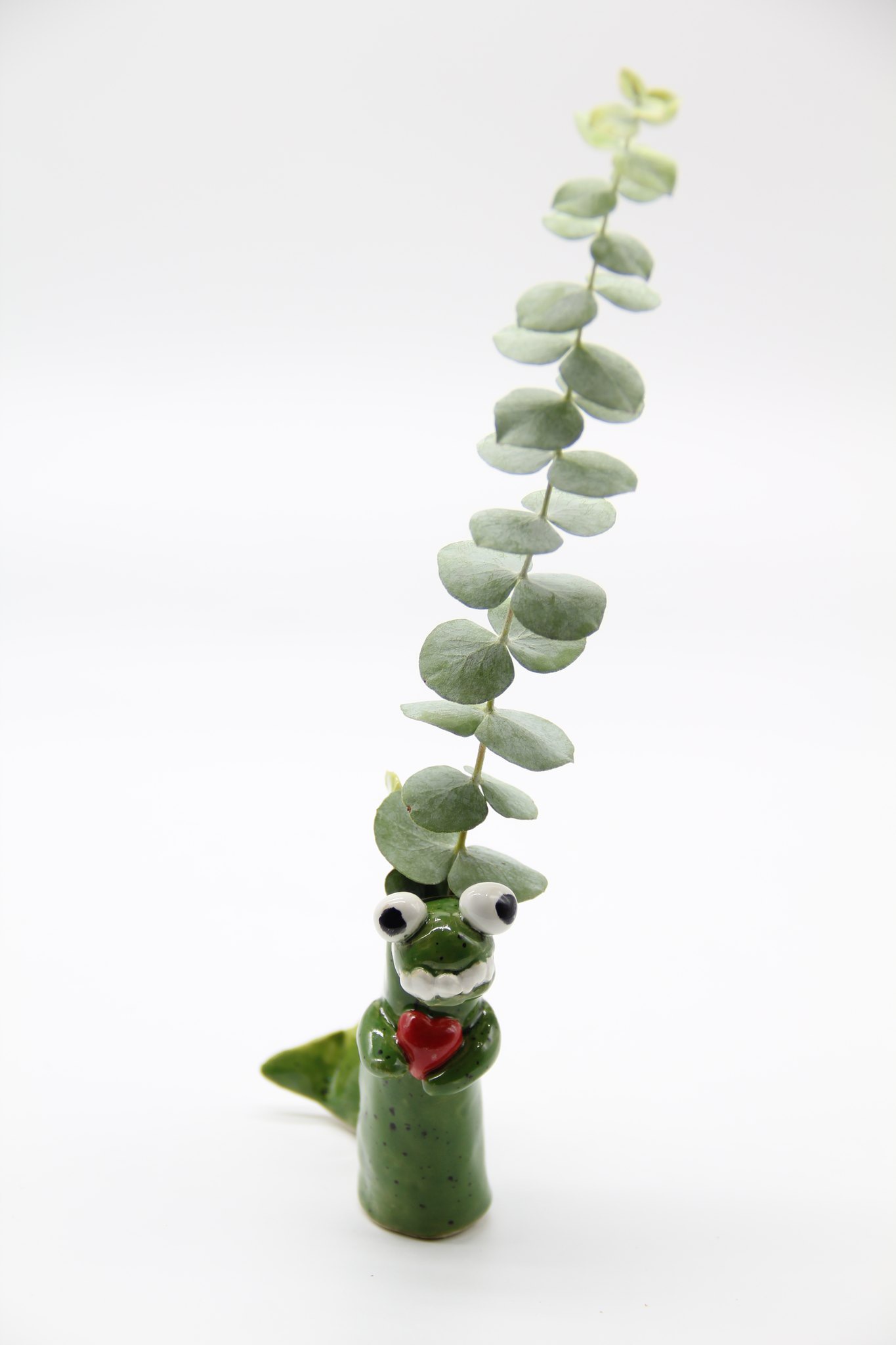 Green toy crocodile with googly eyes and a red tongue, holding a tall eucalyptus branch with round leaves.