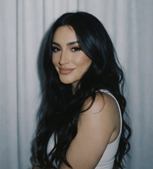 Woman with long dark hair and makeup, smiling, in front of a light-colored curtain.