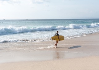 Person carrying a yellow surfboard walking toward the ocean on a sandy beach.