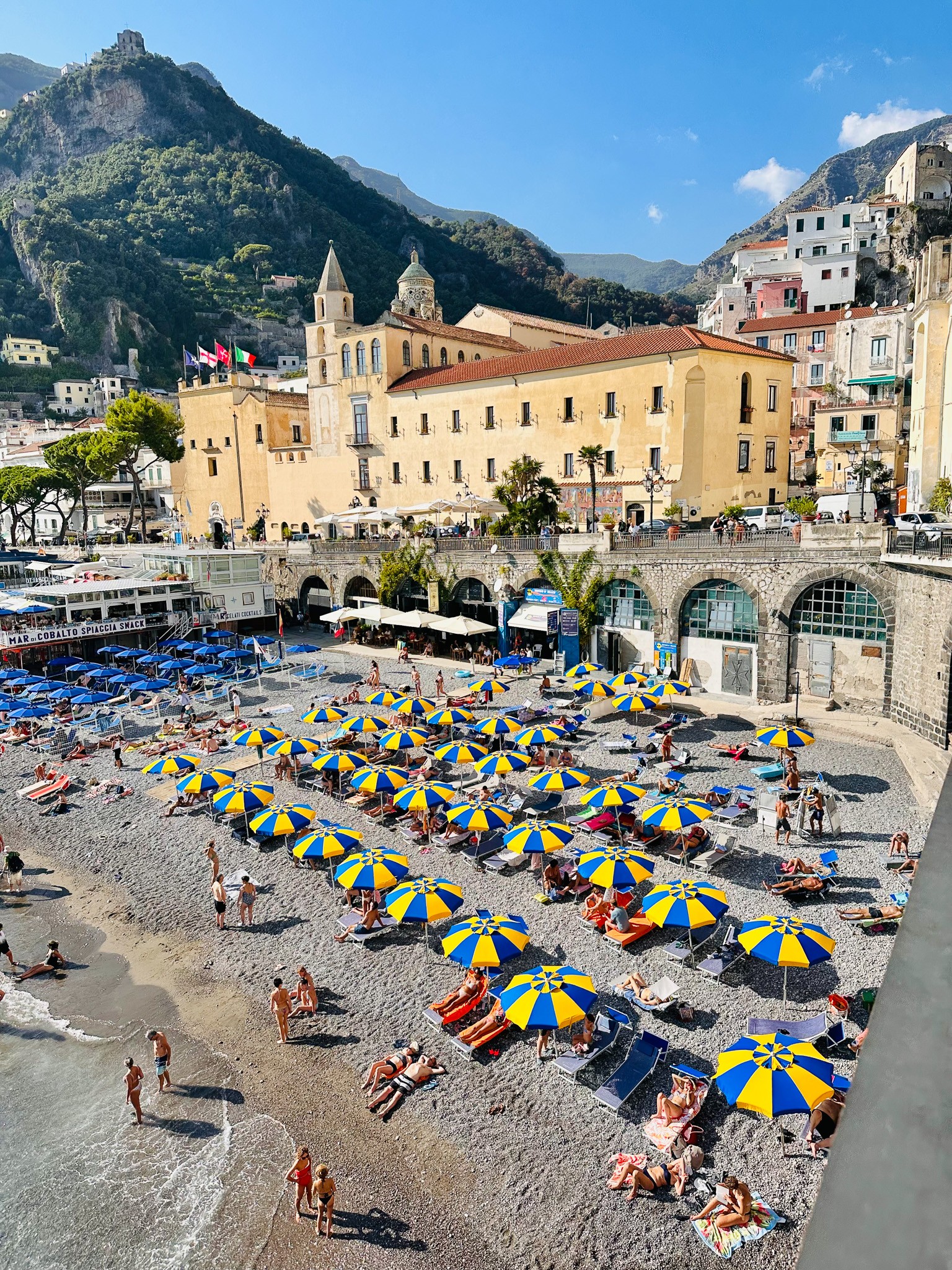 Beach with yellow and blue umbrellas, people relaxing, historic buildings, mountain backdrop, clear sky.