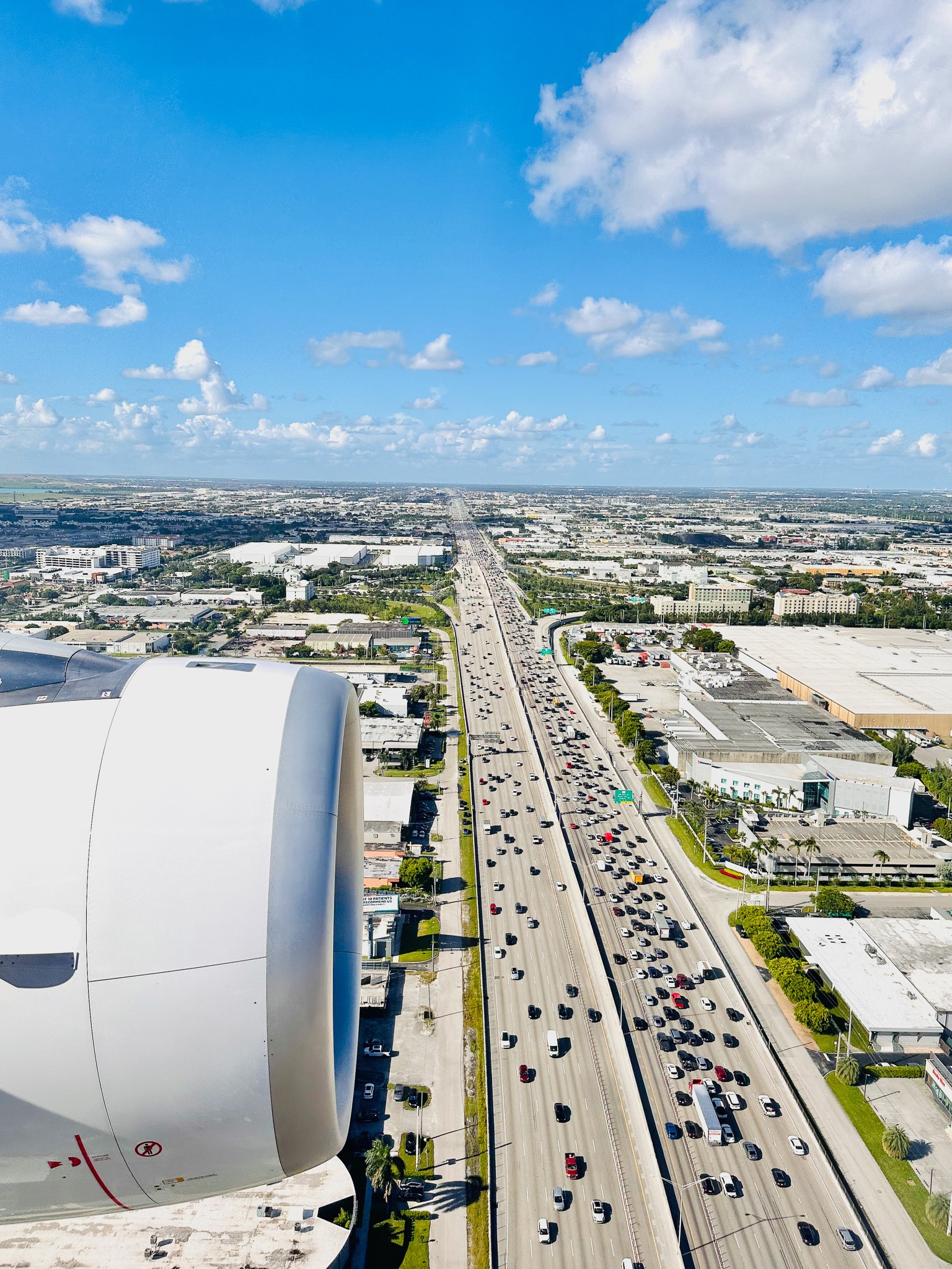 View from an airplane window showing a highway with cars, buildings, and a blue sky with clouds.