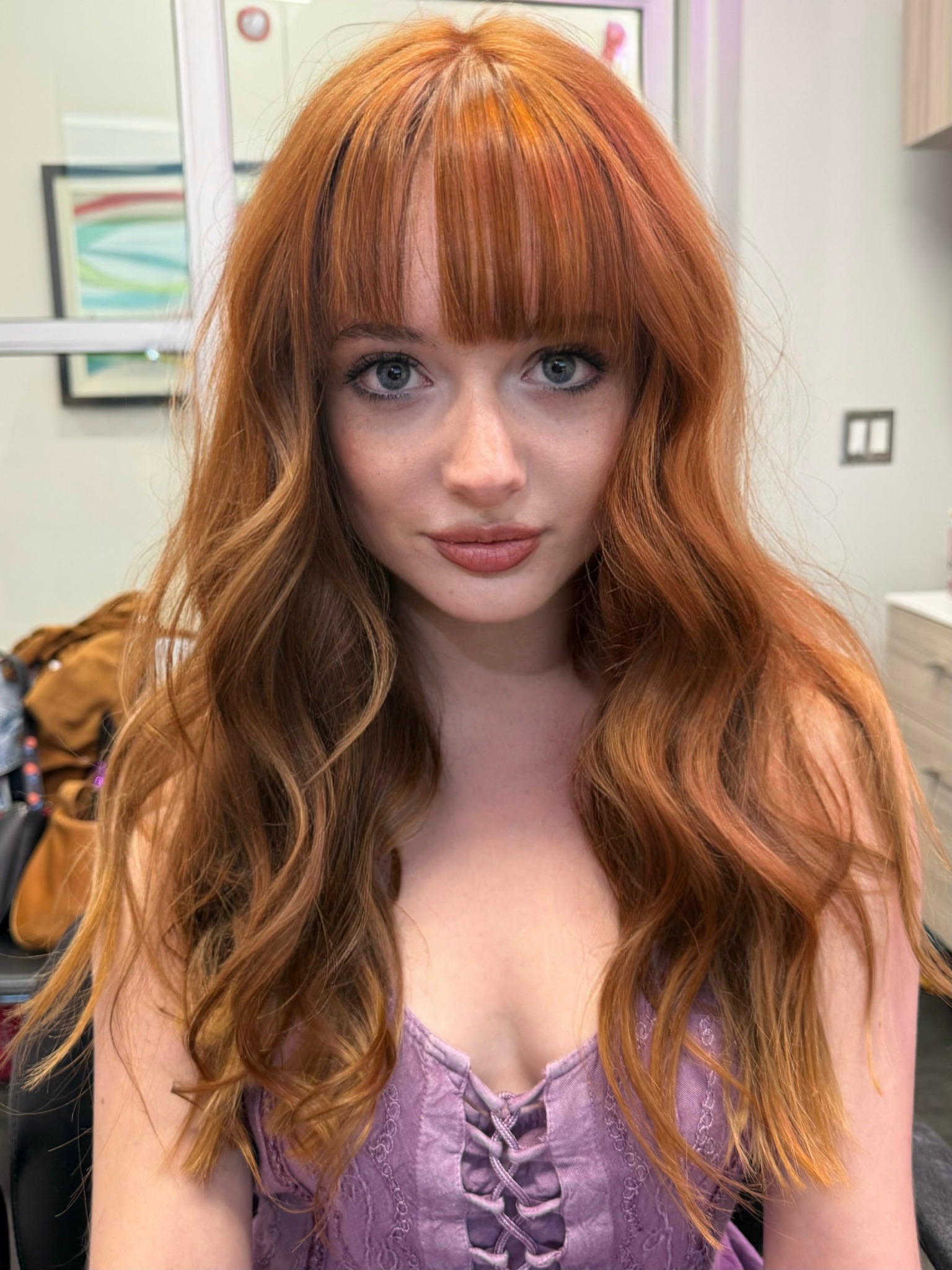 Young woman with long wavy red hair and bangs, wearing a purple top, indoors.