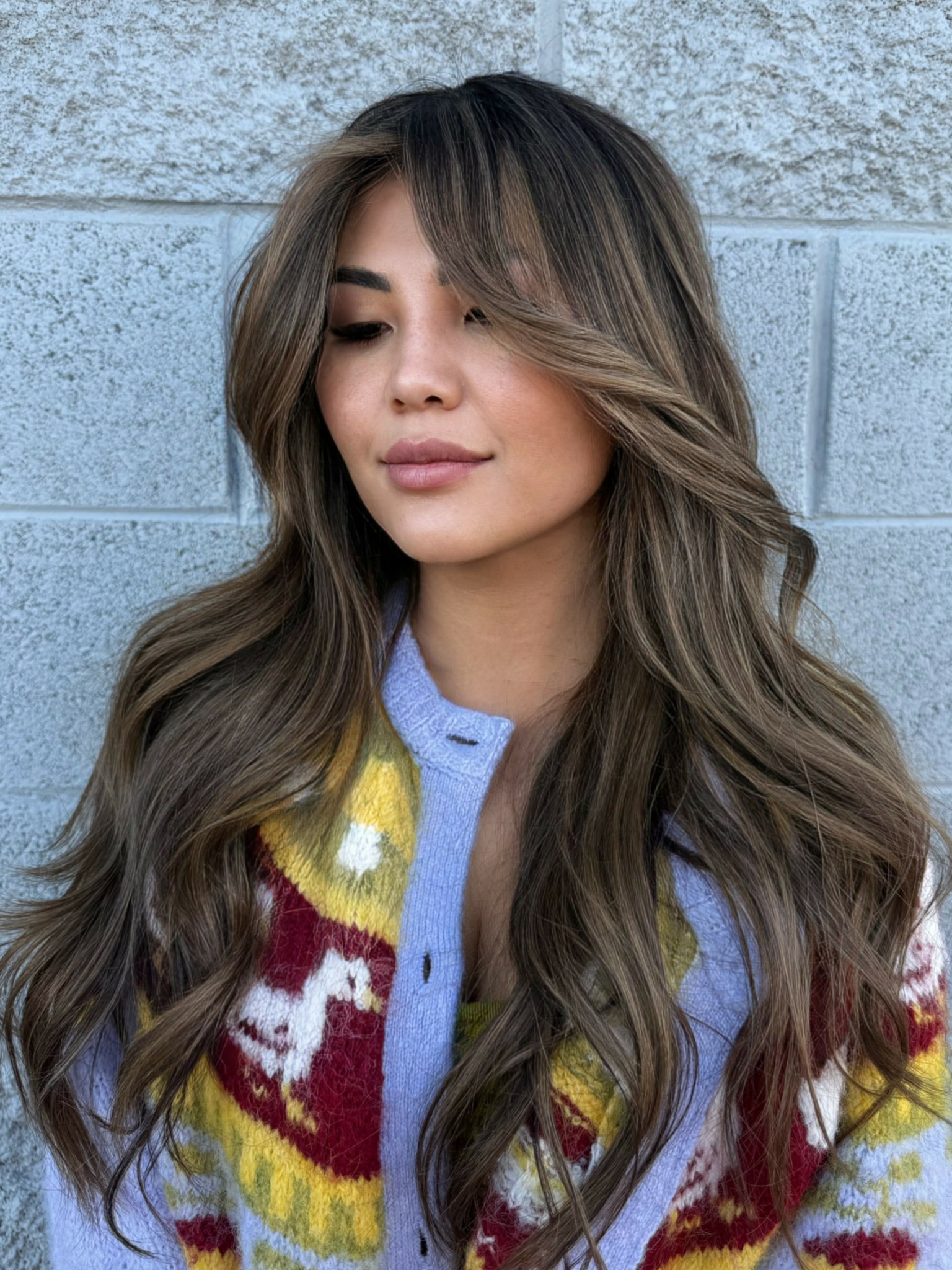 Young woman with long wavy hair and light makeup, standing against a textured gray wall.