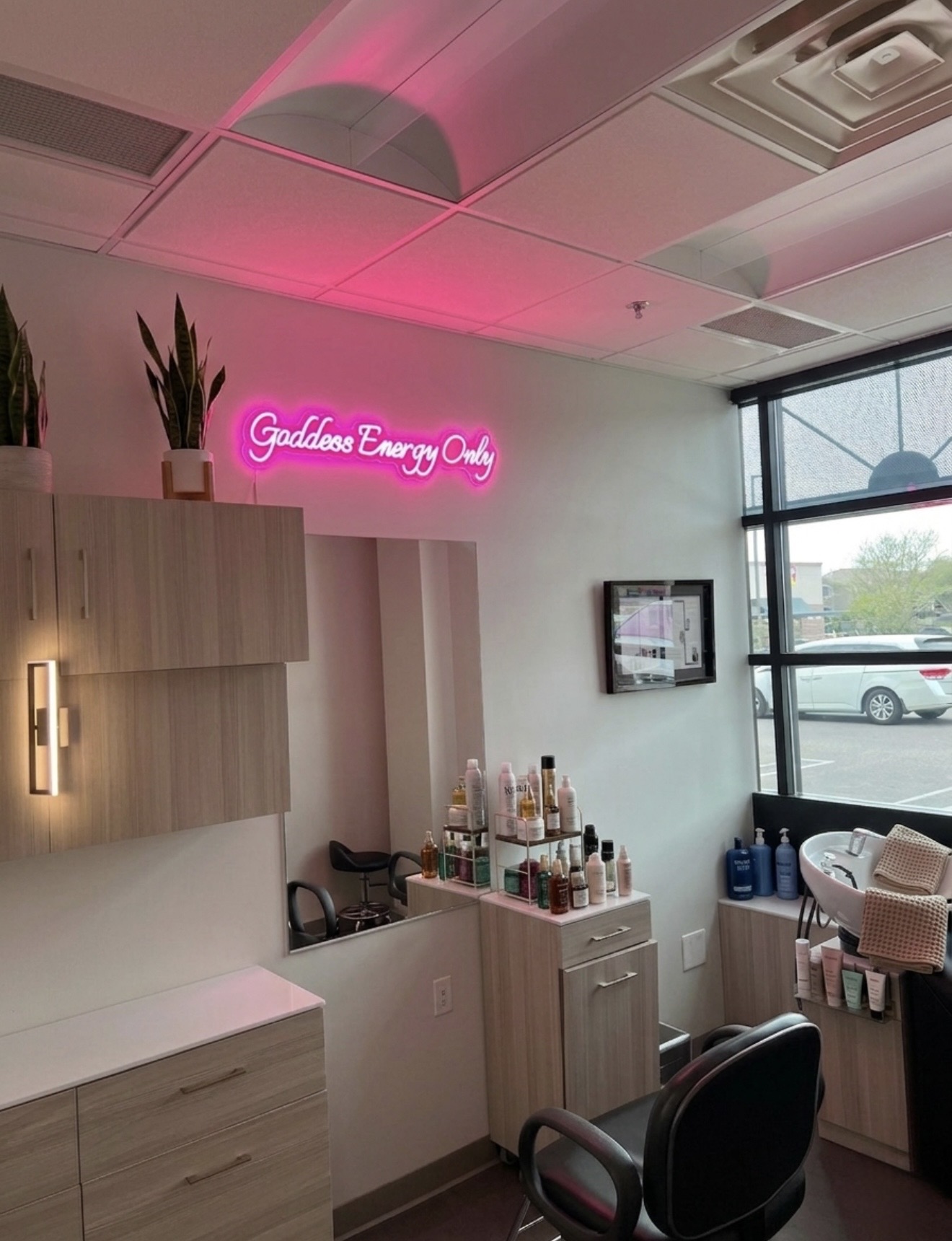 Interior of a salon with a pink neon sign reading 'Goddess Energy Only' on the wall, shelves with products, and a large window.