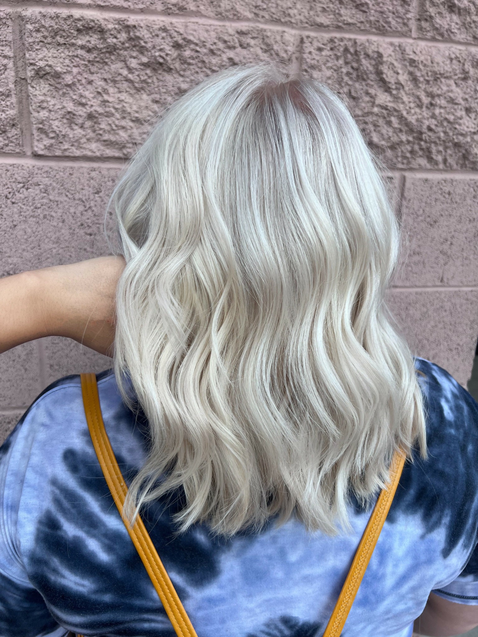 Back of a person with wavy, platinum blonde hair, wearing a blue tie-dye shirt and carrying a yellow strap bag.