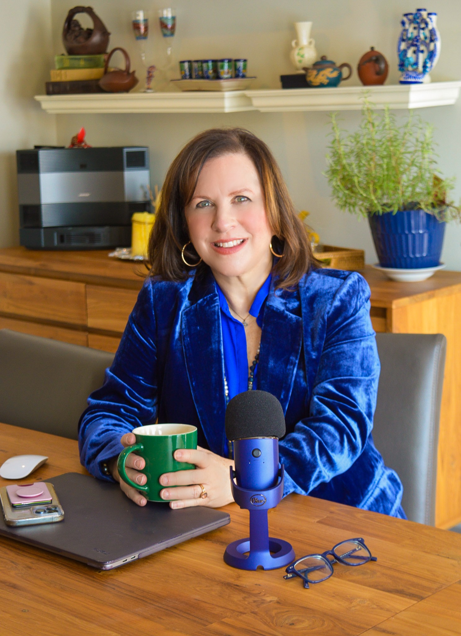 Woman with shoulder-length brown hair, wearing a blue velvet jacket, sitting at a table with a green mug and microphone.