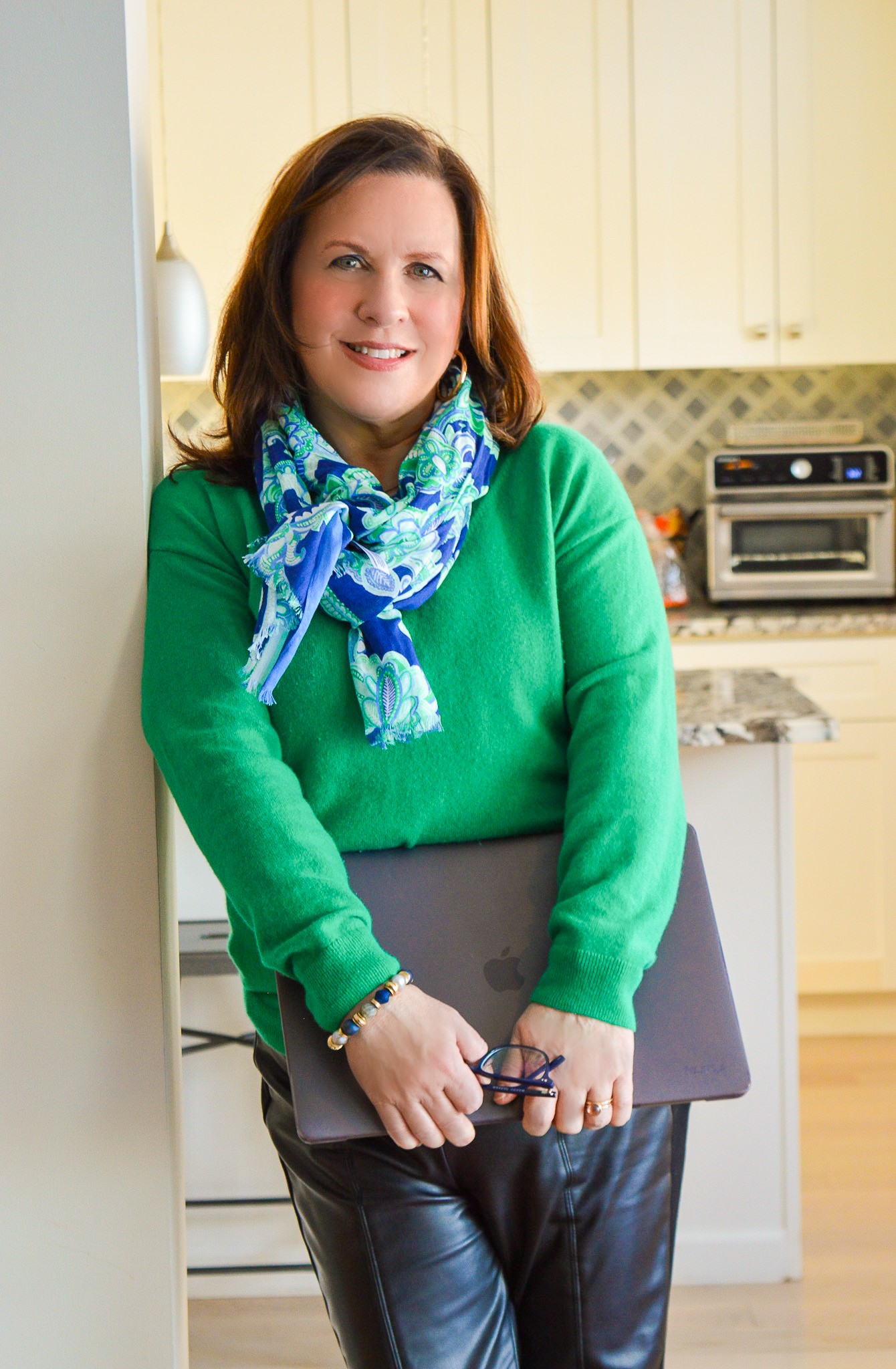 Woman with brown hair wearing a green sweater and blue scarf, holding a laptop, standing in a kitchen.