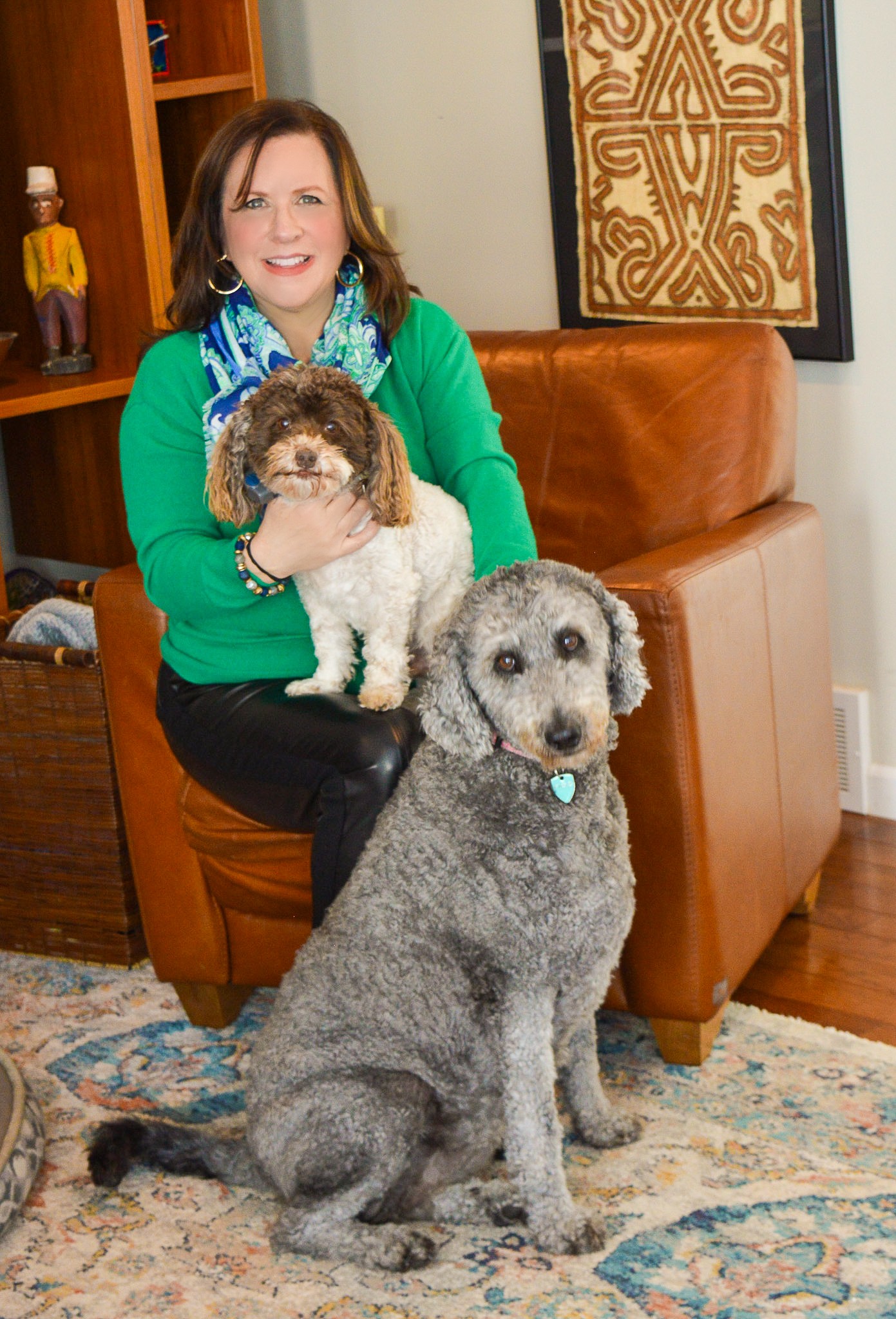 Woman sitting on a sofa holding a small dog, with a large dog sitting on the floor nearby in a living room.