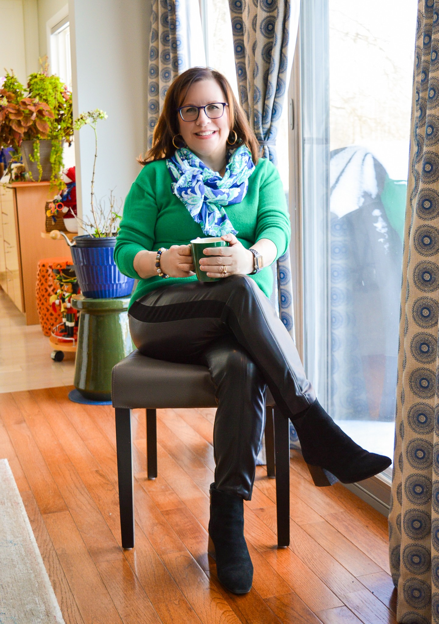 Woman sitting on chair holding a phone, smiling, in a room with curtains and plants.