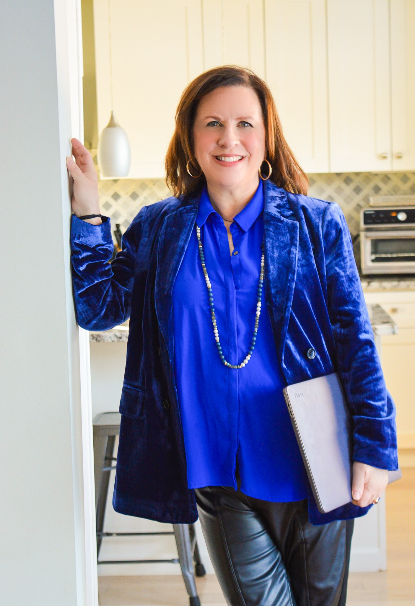 Woman with shoulder-length hair smiling, holding a folder, standing in a kitchen with cream cabinets and a microwave.