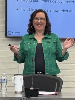 Woman with glasses and dark curly hair smiling, wearing a green jacket, standing in front of a presentation screen.