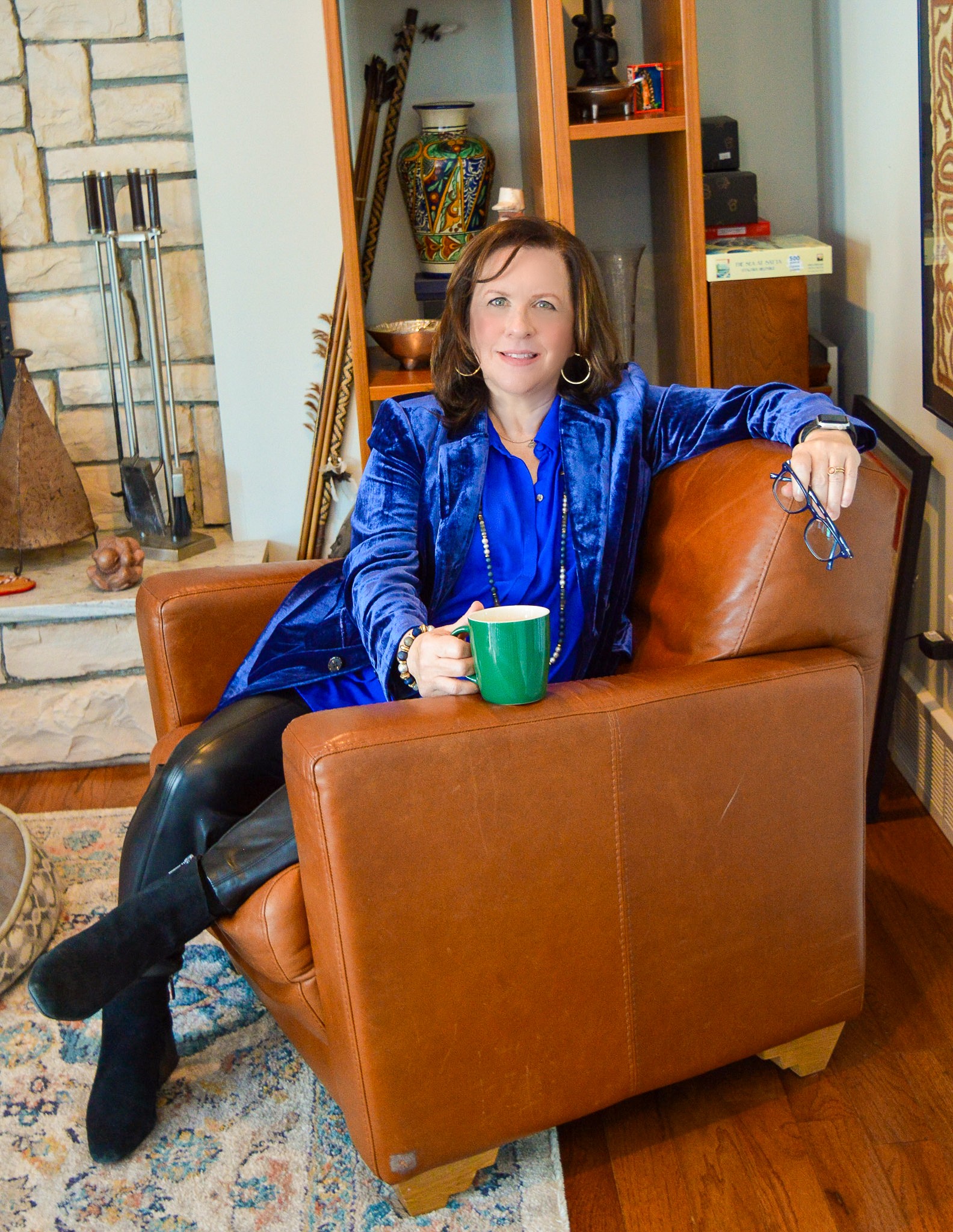 Woman sitting on a brown armchair holding a green mug, smiling, in a cozy room with bookshelves and a fireplace.