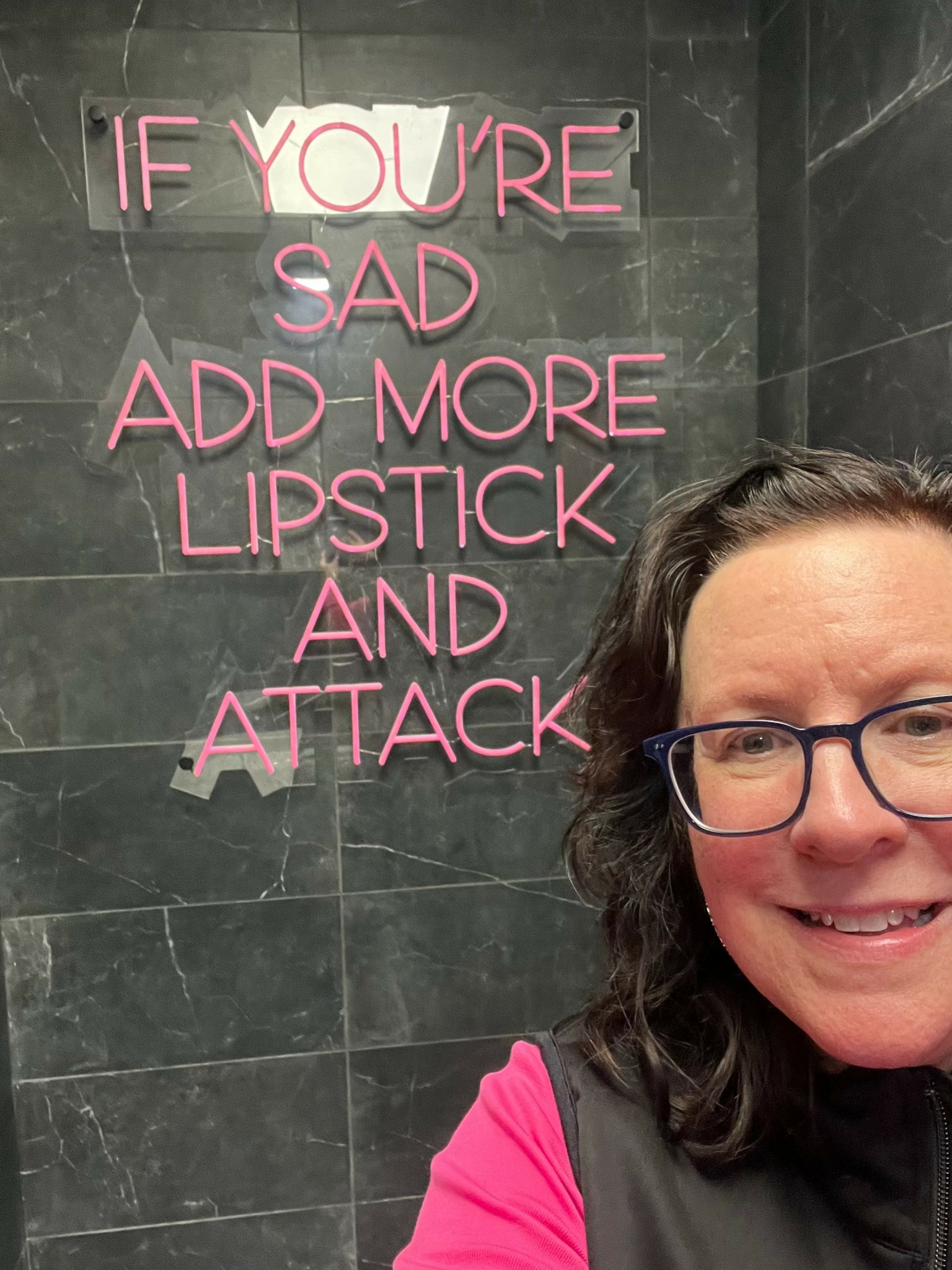 Part of a woman with glasses and dark hair smiling, with a neon sign on a dark wall behind her.