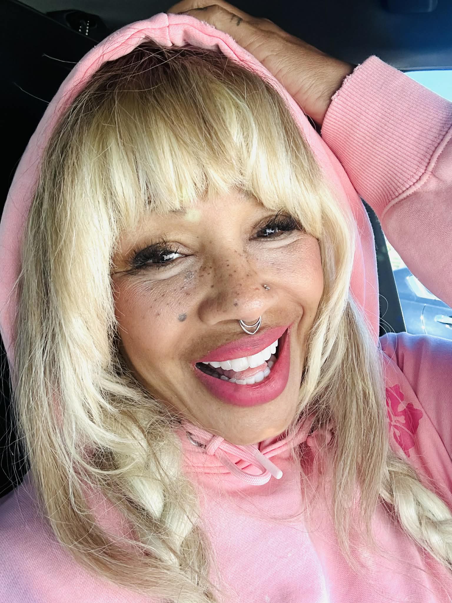 Woman with blonde hair, smiling, wearing pink hoodie, in car interior.