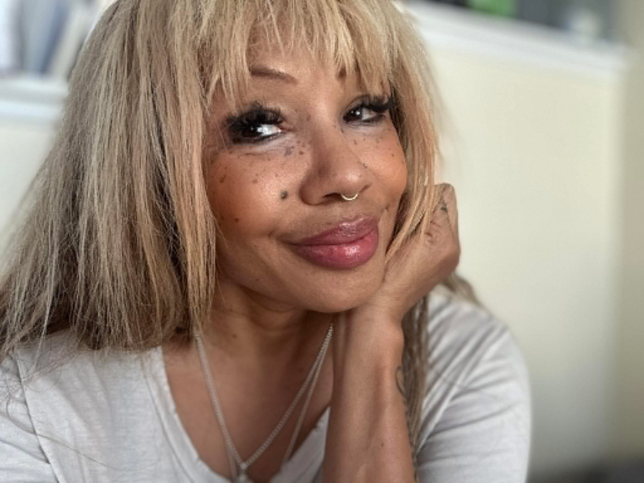 Woman with blonde hair, freckles, and a nose ring, smiling and resting her face on her hand.