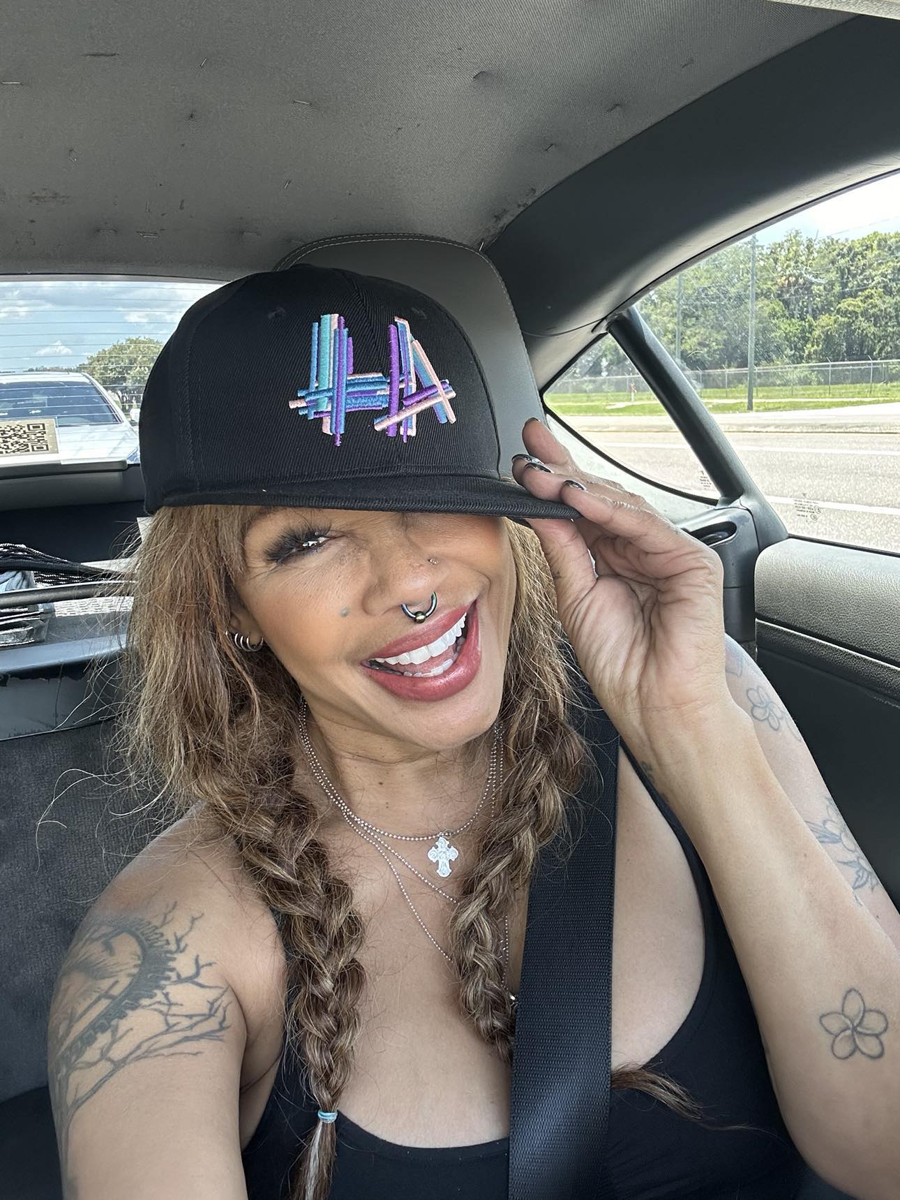 Woman with braids smiling, wearing a black cap with colorful design, inside a car, holding the cap's brim.