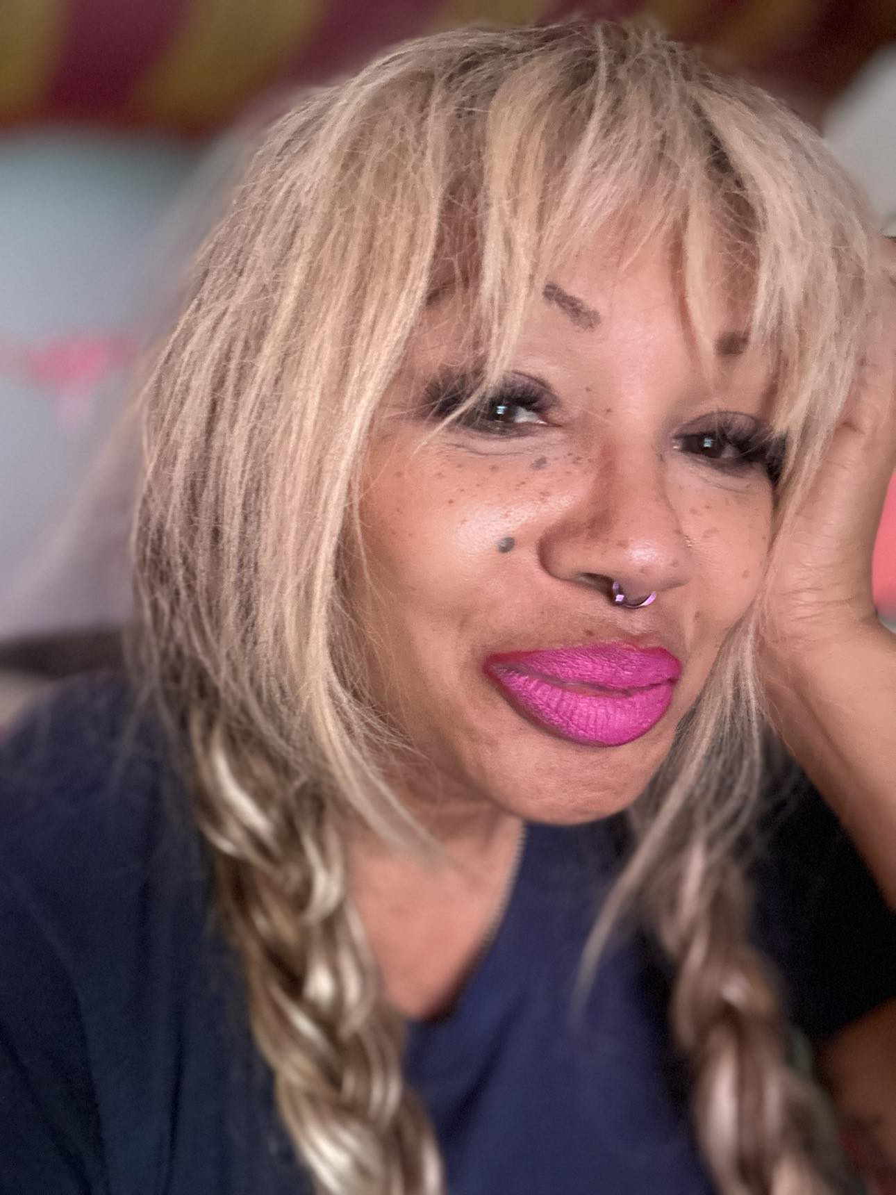 Close-up of a woman with blonde hair, pink lipstick, and facial piercings, resting her face on her hand.