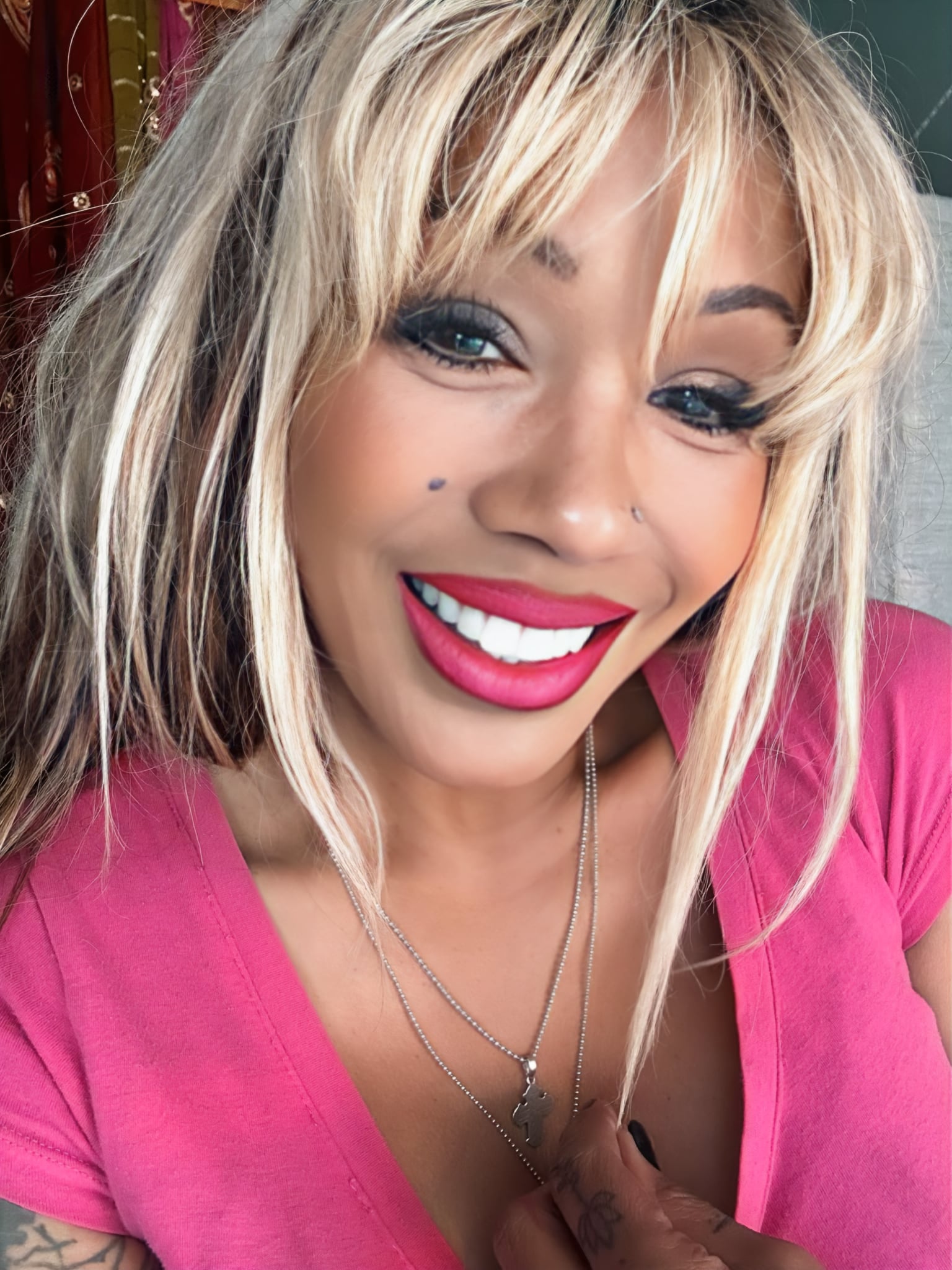 Close-up of a smiling woman with blonde hair, makeup, pink lipstick, wearing a pink top and layered necklaces.