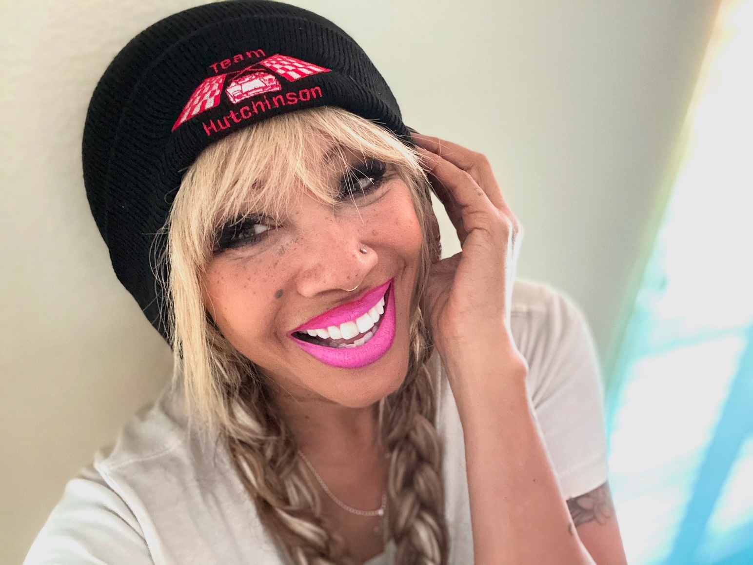 Woman with blonde hair, wearing a black beanie, smiling, touching her face, in a light-colored room.