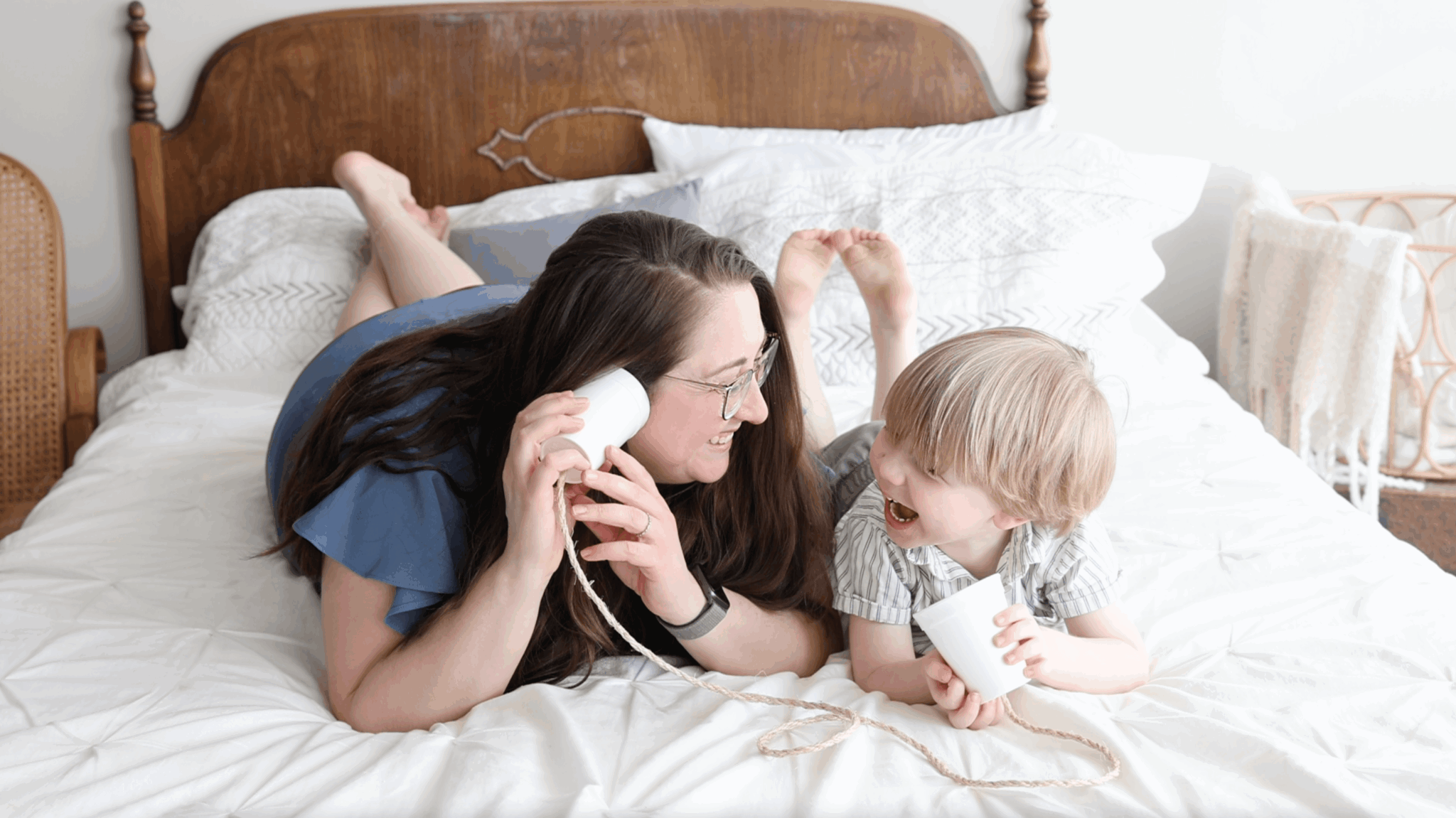 Woman and child lying on bed, talking and smiling, with a phone and a toy, in a bedroom.