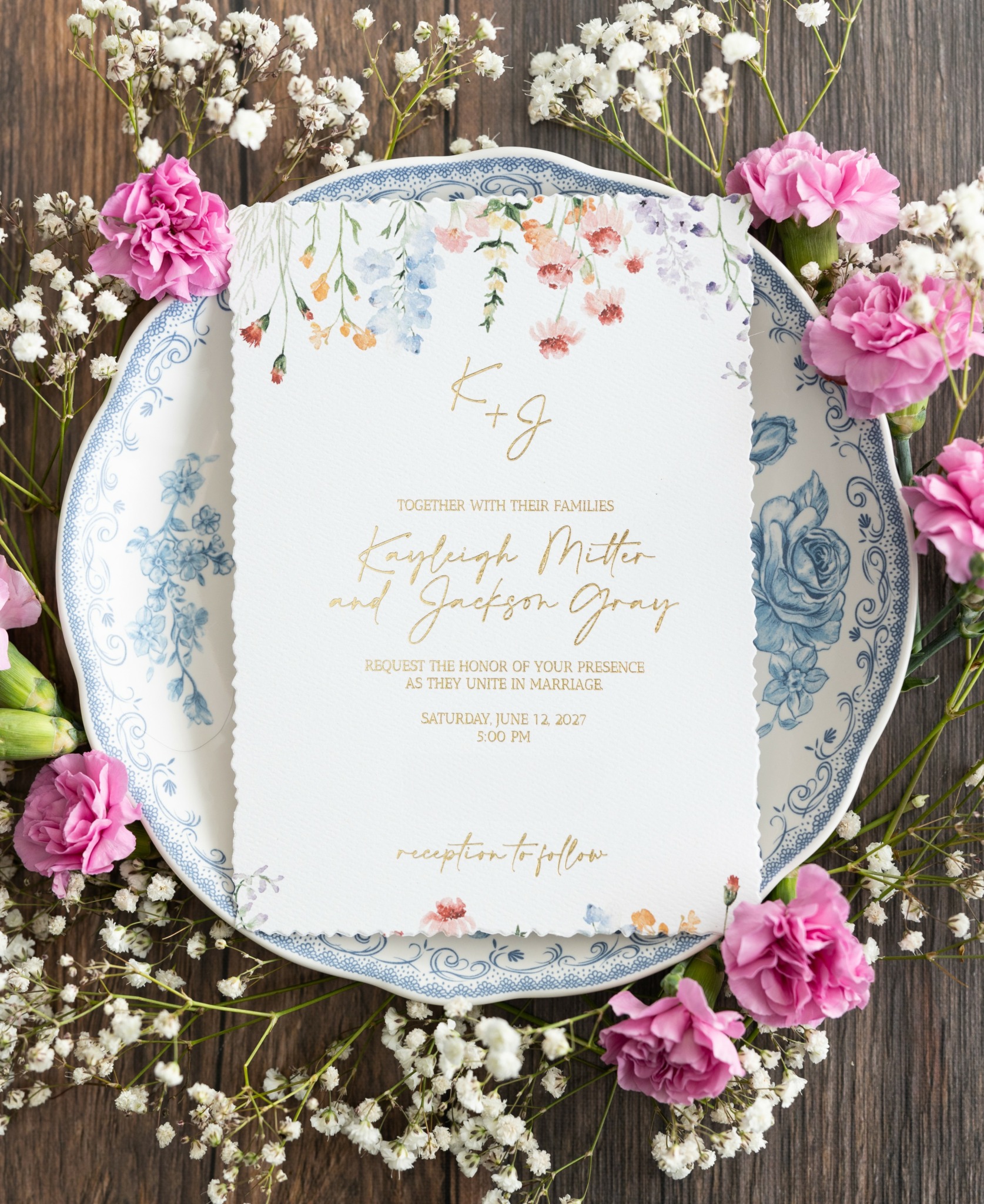 Invitation card on decorative plate surrounded by pink and white flowers on wooden surface.