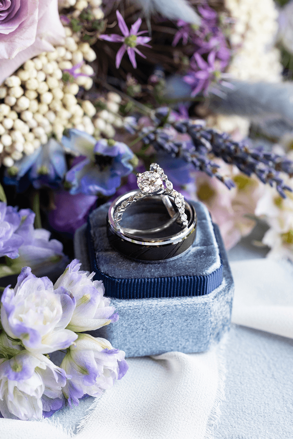 Diamond engagement ring on a blue velvet box surrounded by purple, white, and lavender flowers.
