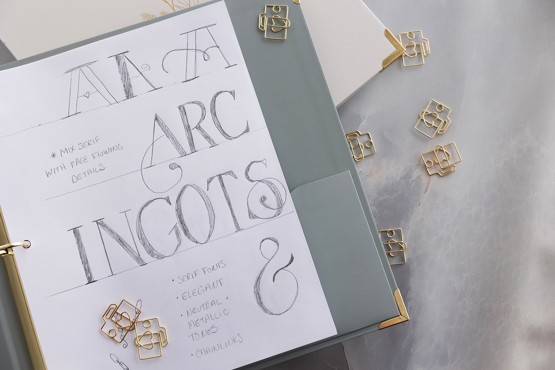 Open notebook with handwritten words 'A, A, ARC, INCOTES' and decorative gold clips on a light surface.