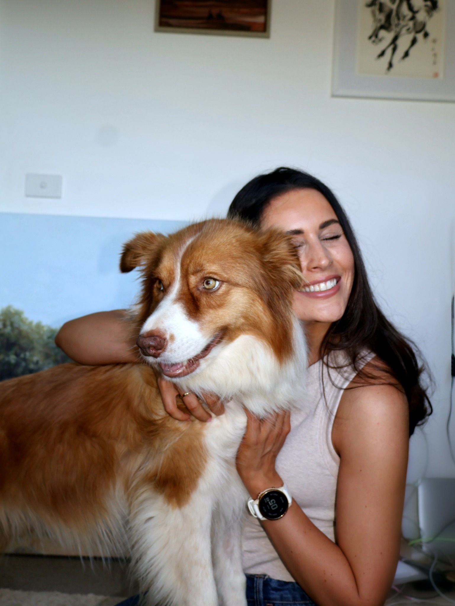 Woman smiling with a large, brown and white dog indoors, wall with framed pictures behind them.