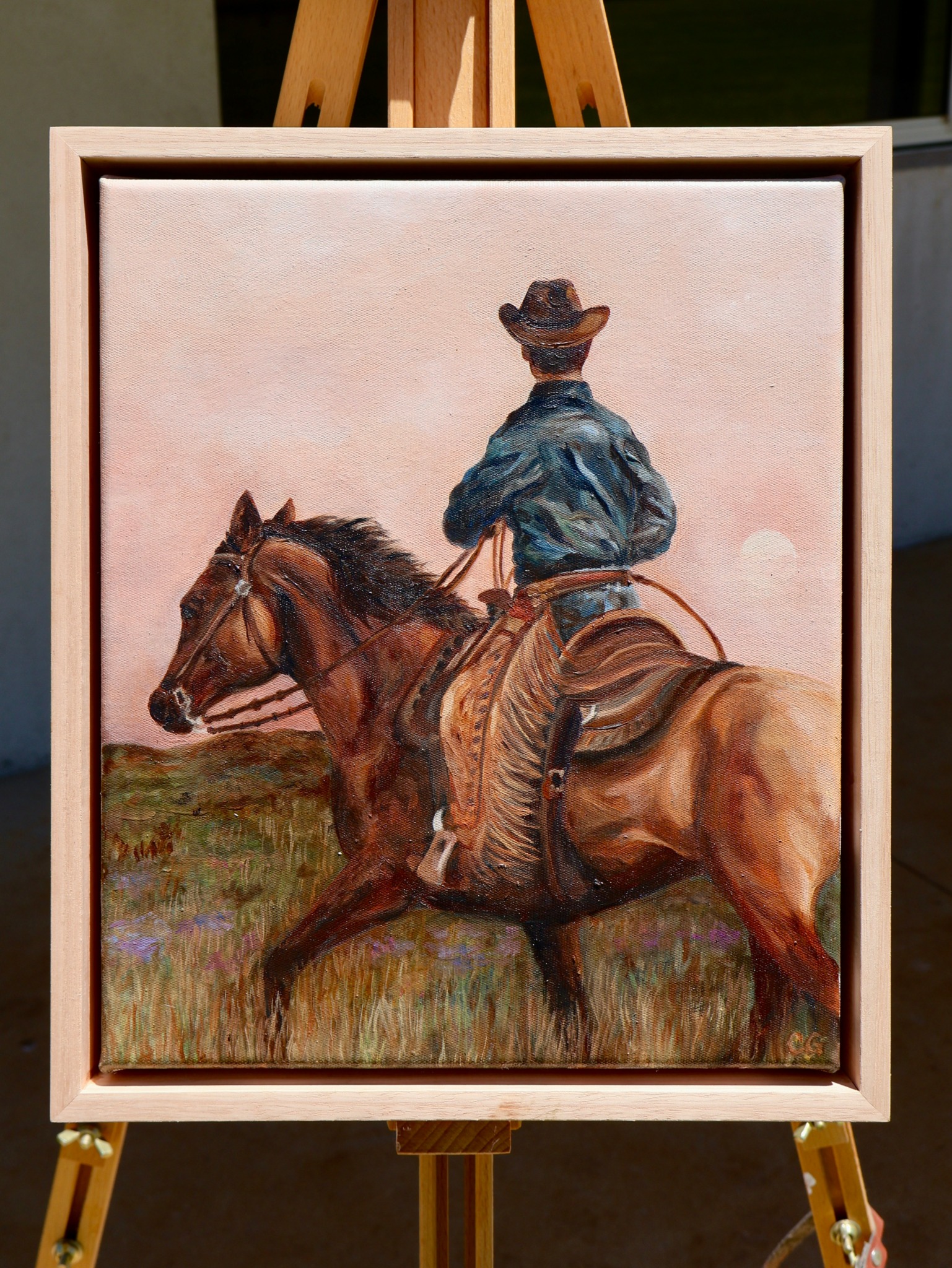 Person wearing a hat riding a brown horse in an open field, with a pinkish sky background.