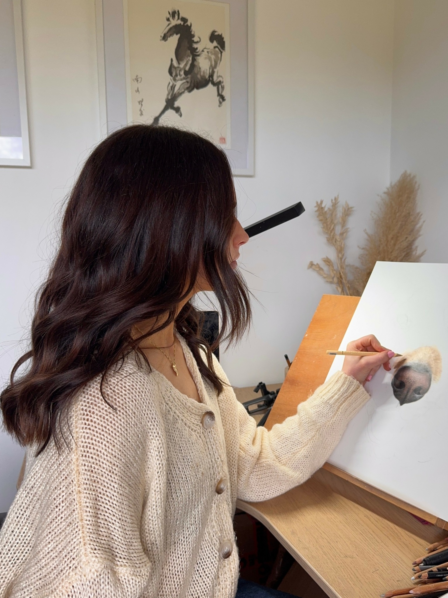 Person with wavy dark hair painting on a white canvas in a room with beige walls and dried plants.