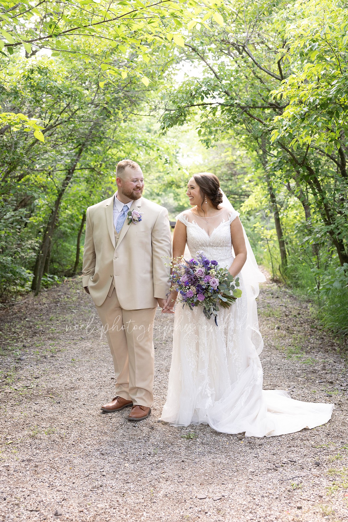 Bride and groom walking on a forest path, surrounded by green trees, holding a bouquet of flowers.