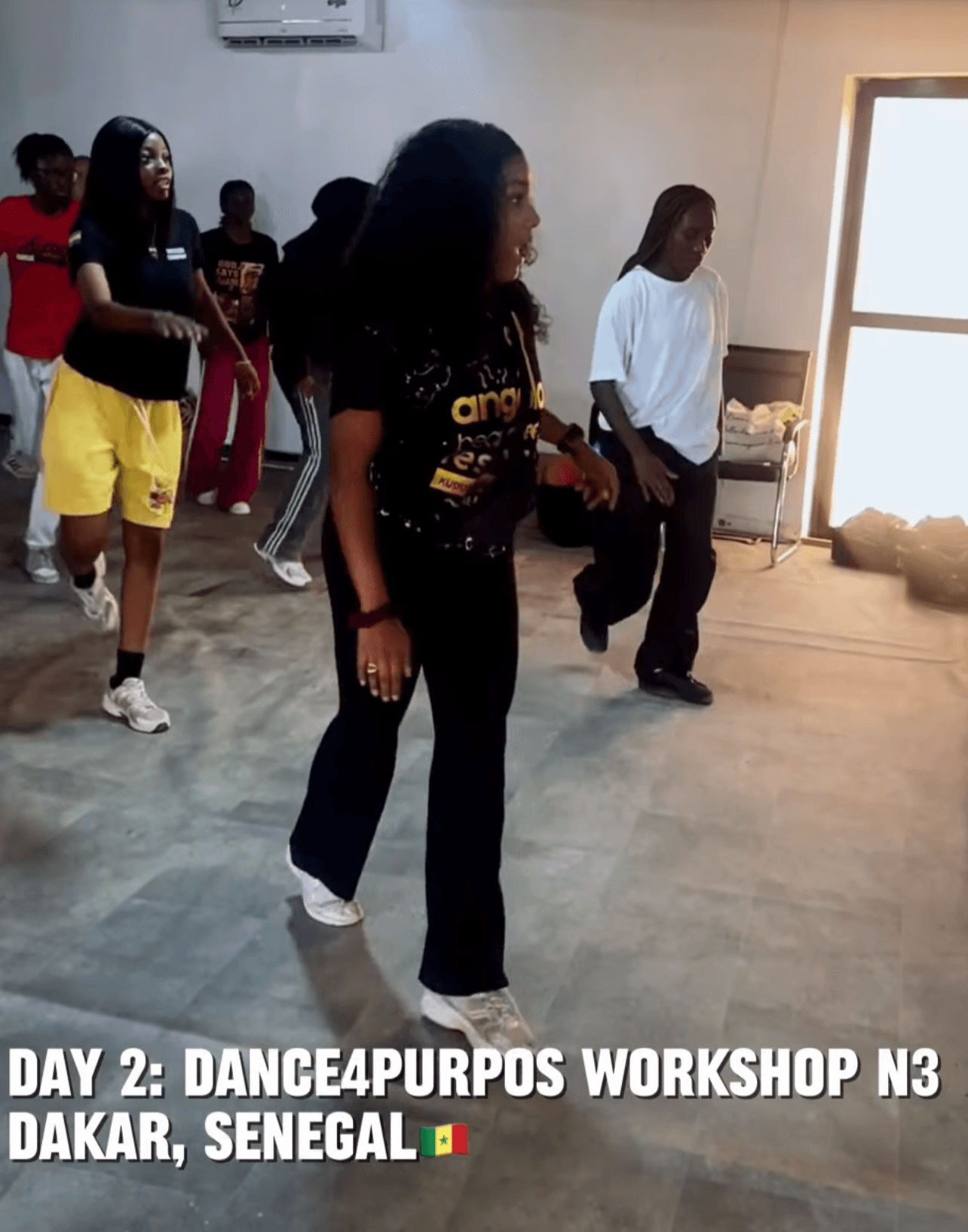 Group of people in a dance workshop room, some dancing, others watching, with a door and window in the background.