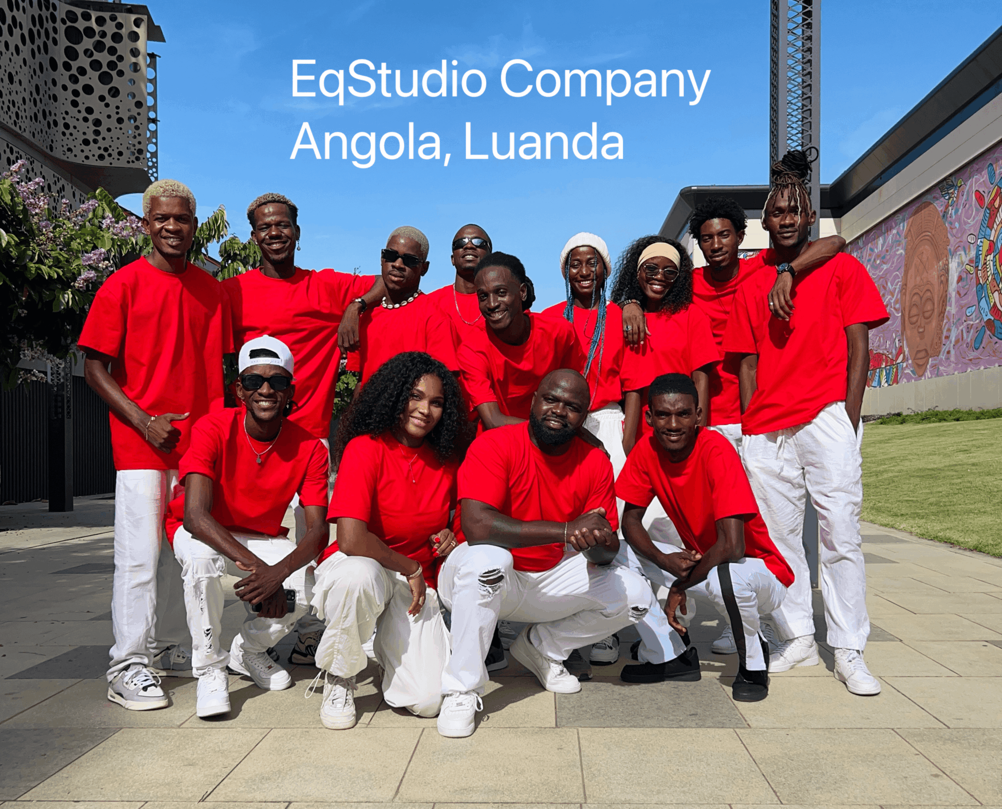 Group of ten people wearing red shirts and white pants outdoors, with a mural and building in background.