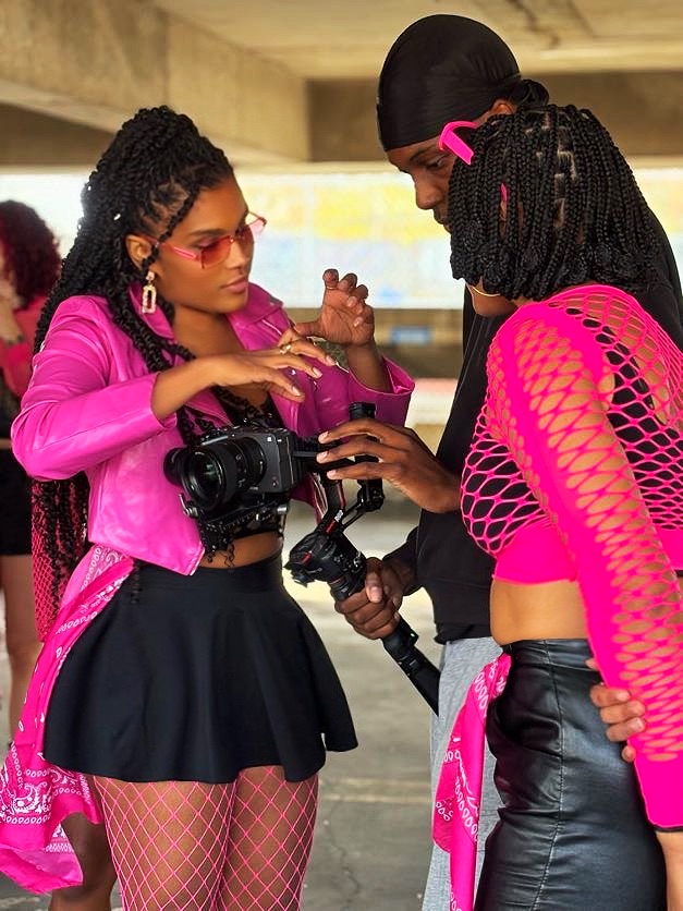 Two young women with cameras and accessories, wearing pink and black outfits, standing indoors and talking.
