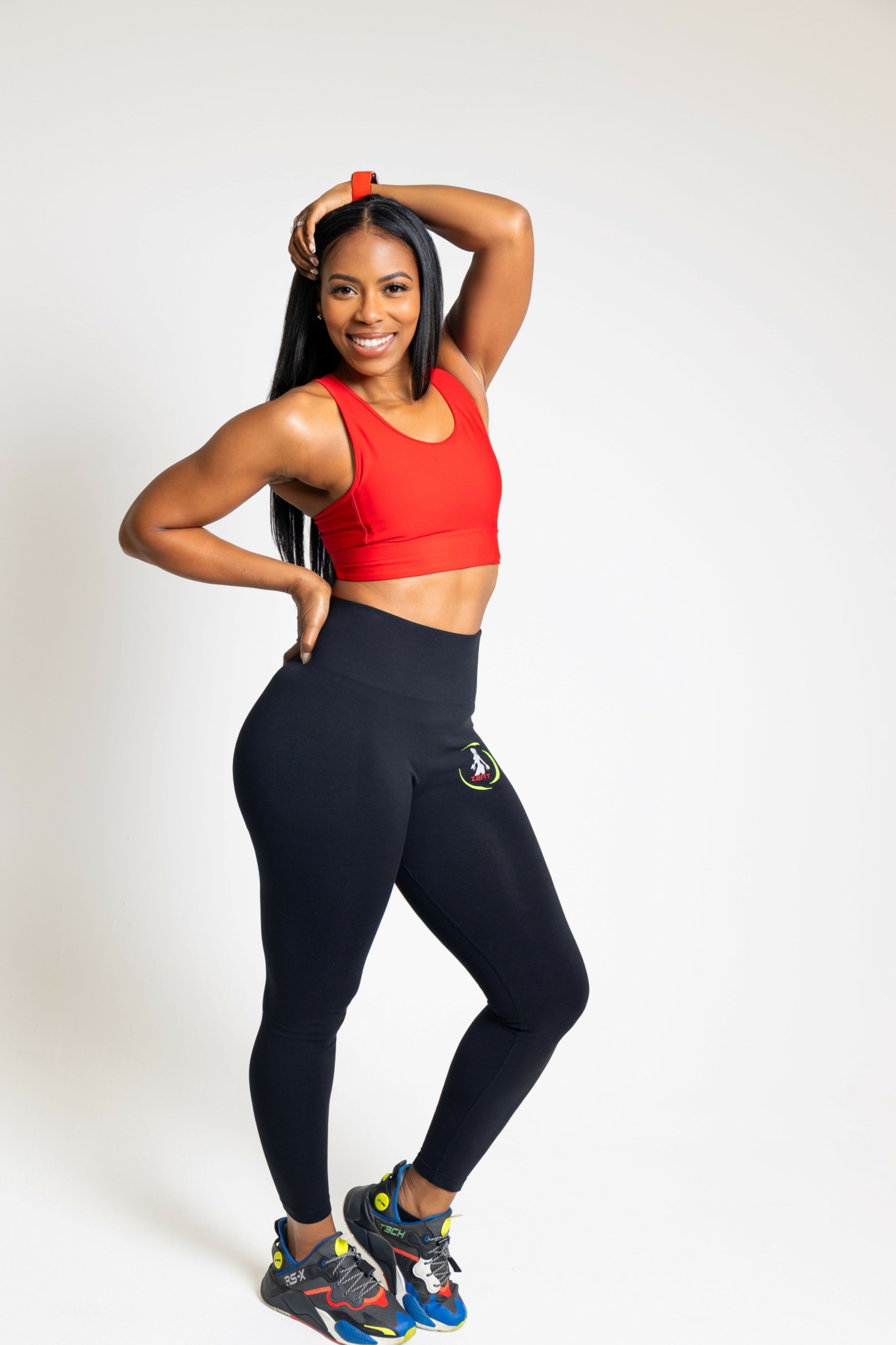 Woman in red sports bra and black leggings smiling with hand on head and hip, standing against white background.