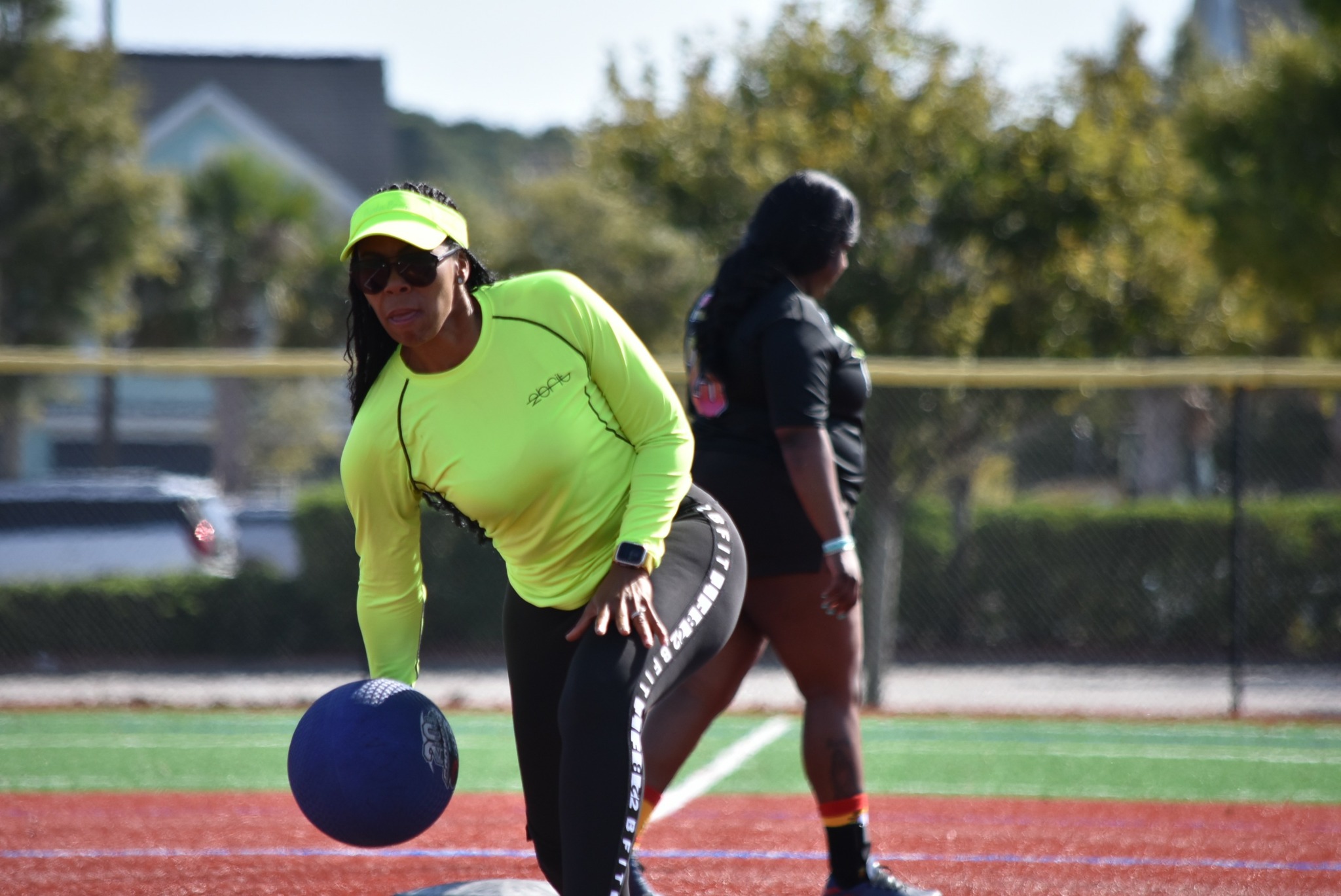 Woman in bright yellow sportswear reaching for a discus on a track field, with another person in the background.