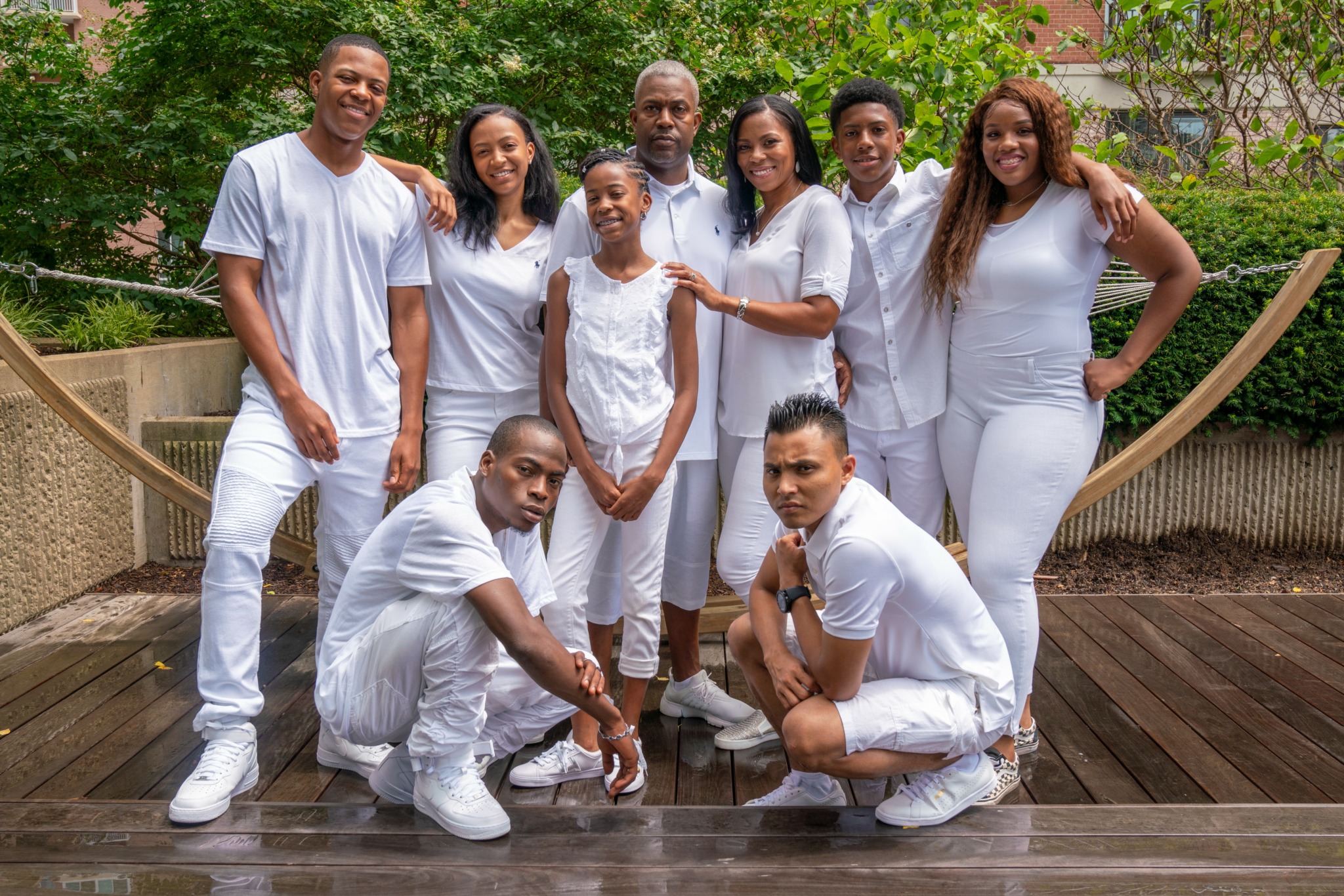 Group of eight people posing outdoors on a wooden deck with greenery in the background.
