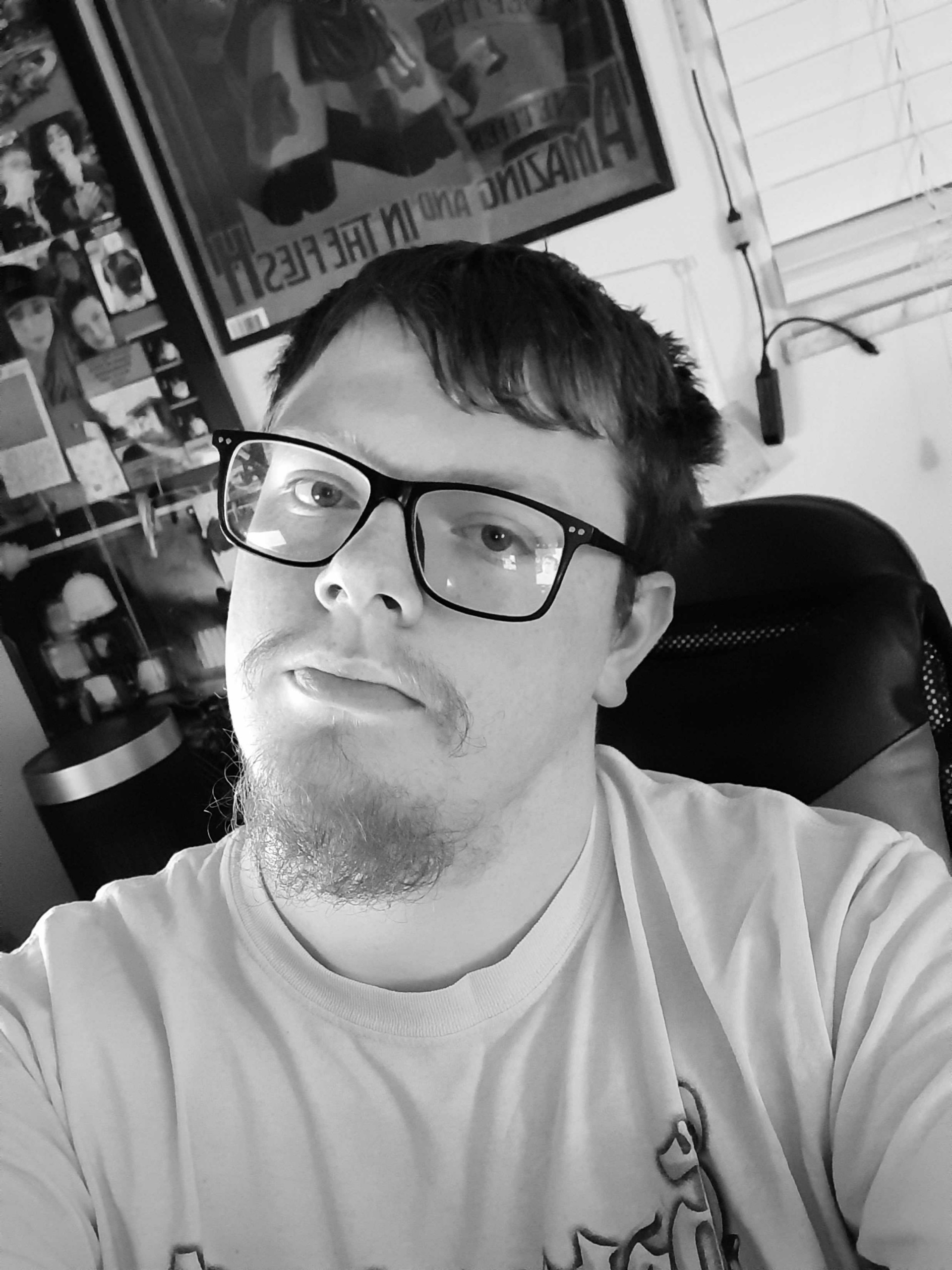 Young man with glasses and short hair taking a selfie indoors, smiling slightly, wearing a light-colored shirt.