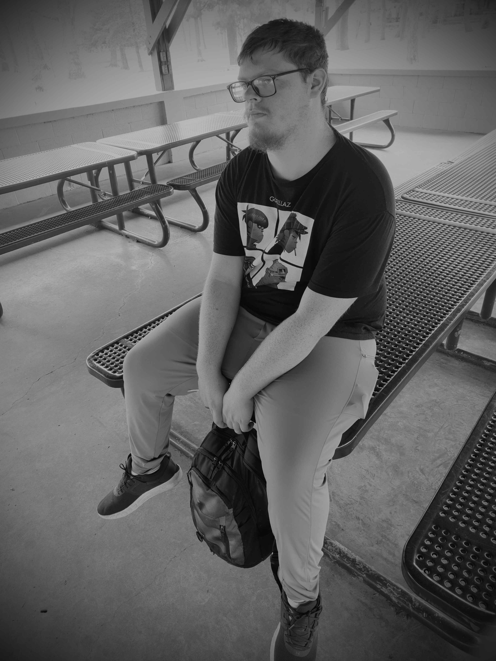 Person sitting on a bench with a backpack, wearing glasses and casual clothing, in an indoor setting.